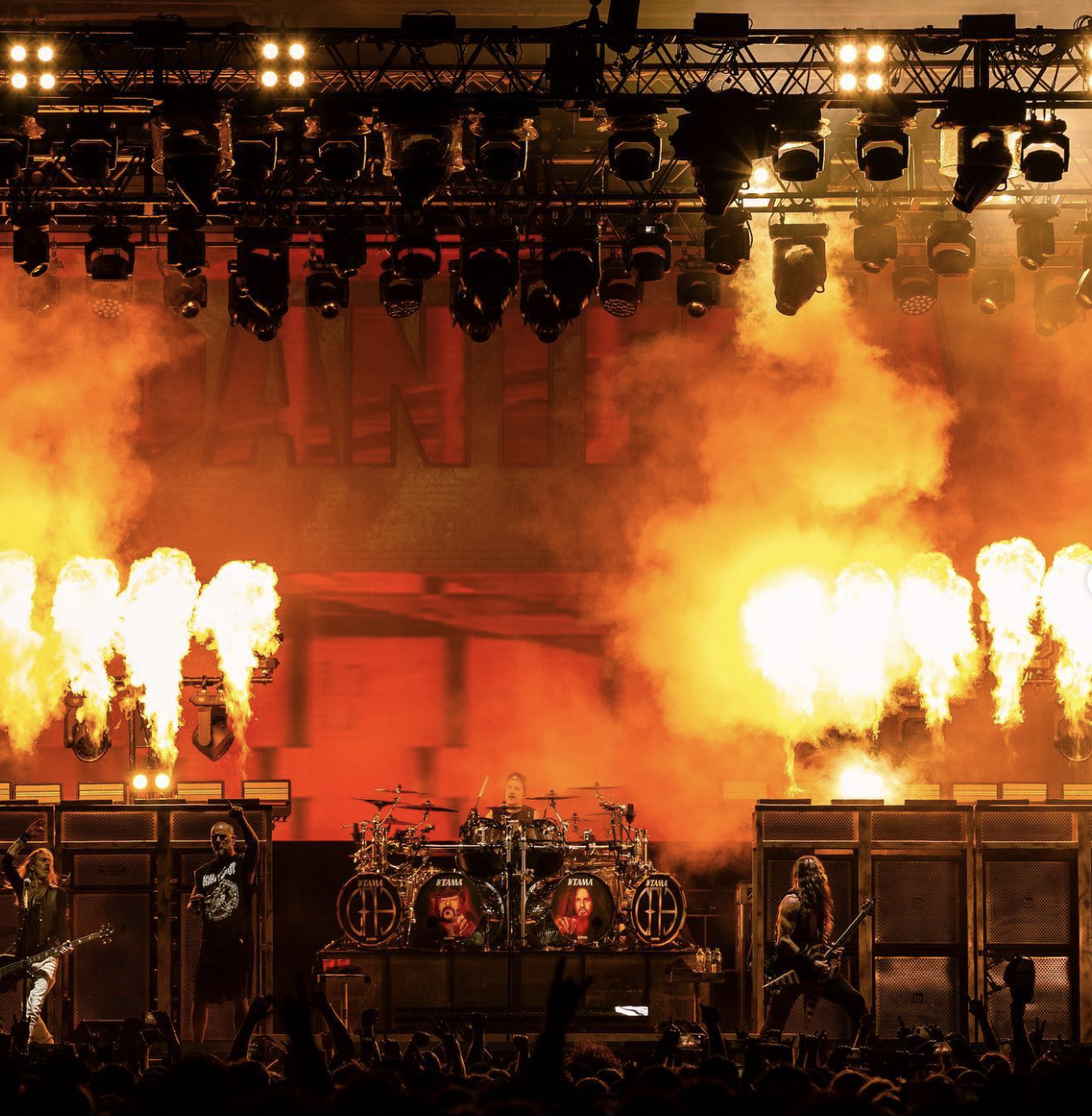 A rock concert stage with a drummer, guitarist, and vocalist, surrounded by pyrotechnics and bright lights, with an audience in the foreground.