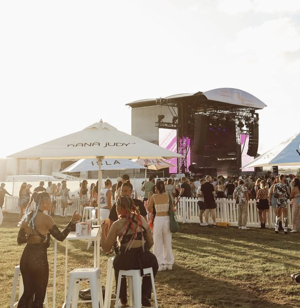 Crowd at an outdoor music festival with stage, large screens, and white tents, some with seating and umbrellas labeled 'NANA JUDY.'