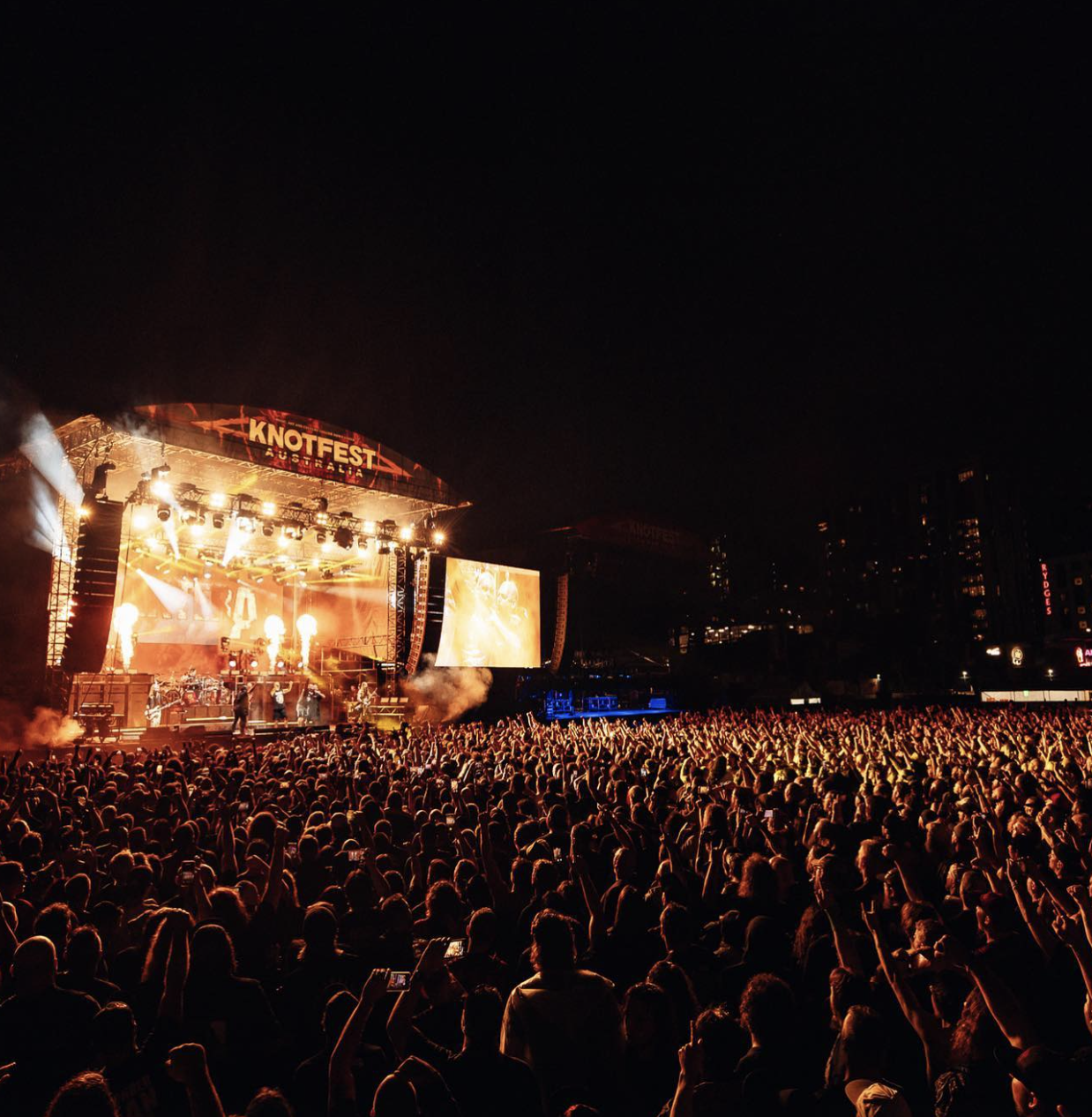 Crowd at a music festival at night with a large stage and bright lights, banners reading 'KNOTEST AUSTRALIA', and screens displaying the performance.