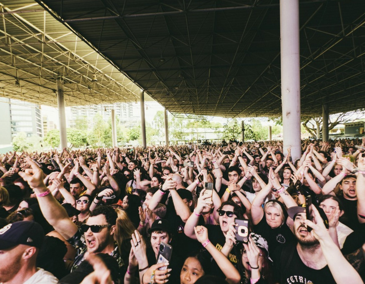 Crowd of concert-goers under a large outdoor roof, raising hands and recording with smartphones.
