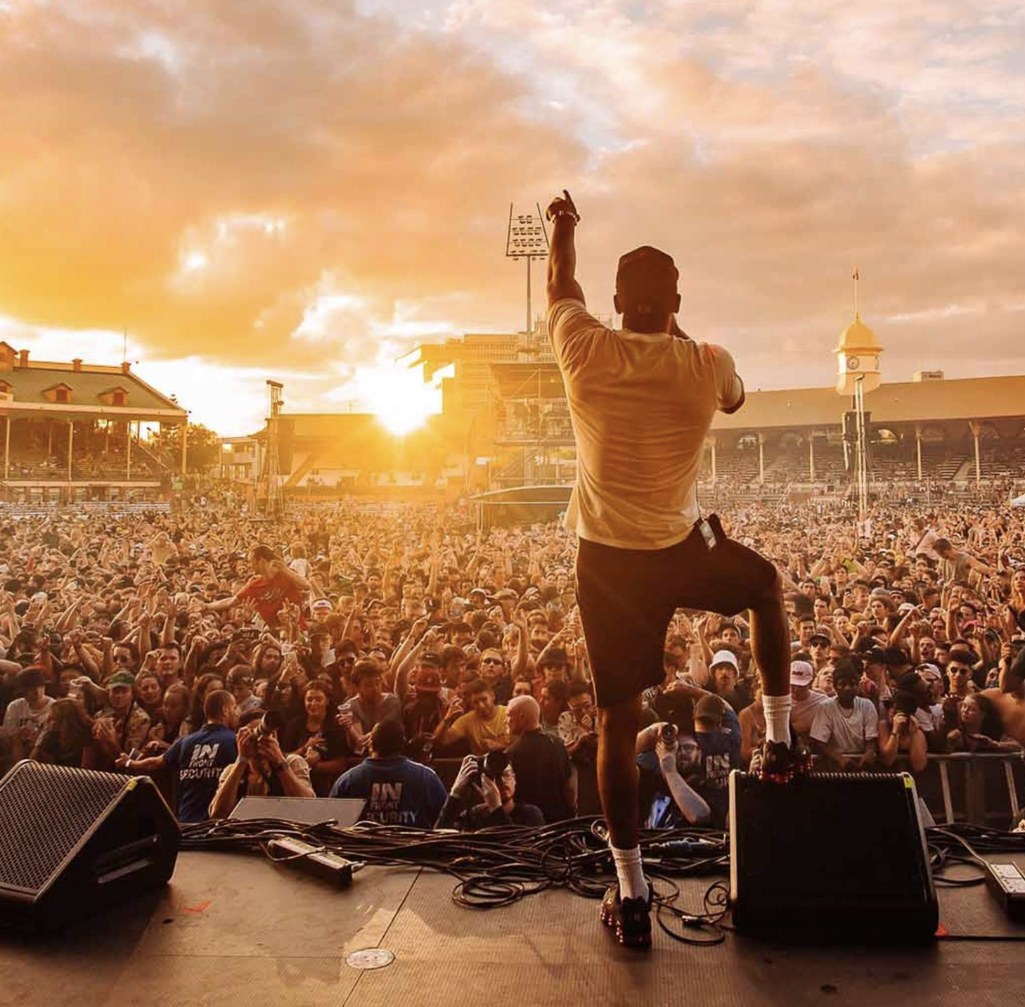 A musician or performer on stage in front of a large crowd at a concert during sunset, with one arm raised and facing away from the camera.