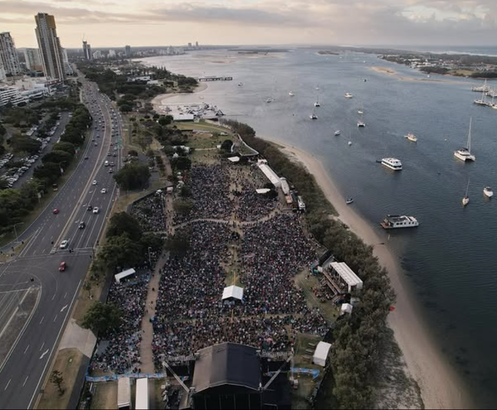 A large outdoor concert by the waterfront with a dense crowd of attendees, boats anchored on the water, high-rise buildings to the left, and a scenic view of the city skyline and water on a cloudy day.