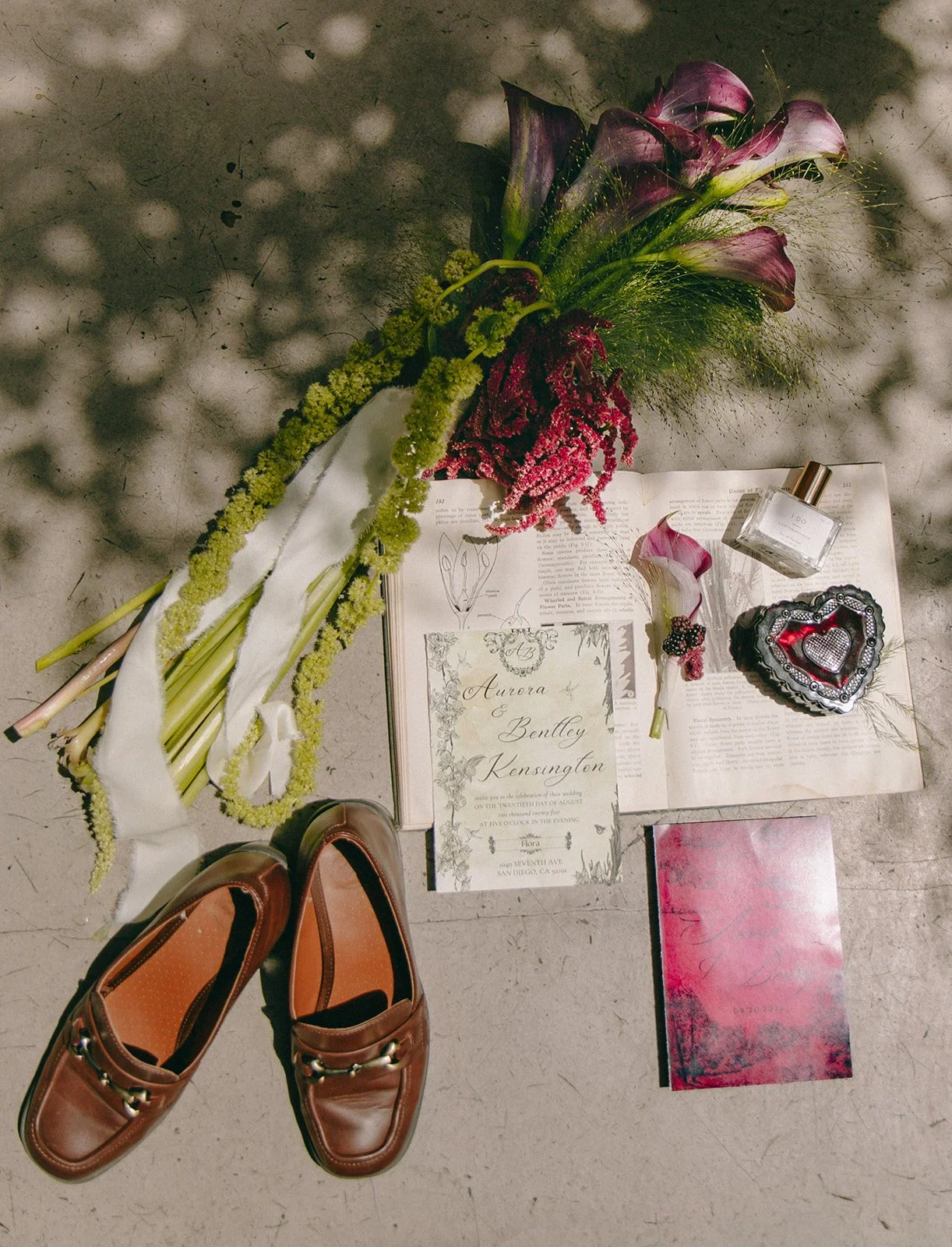 A floral arrangement, a pair of brown loafers, wedding invitation, perfume, decorative box, and small book are laid out on the ground.