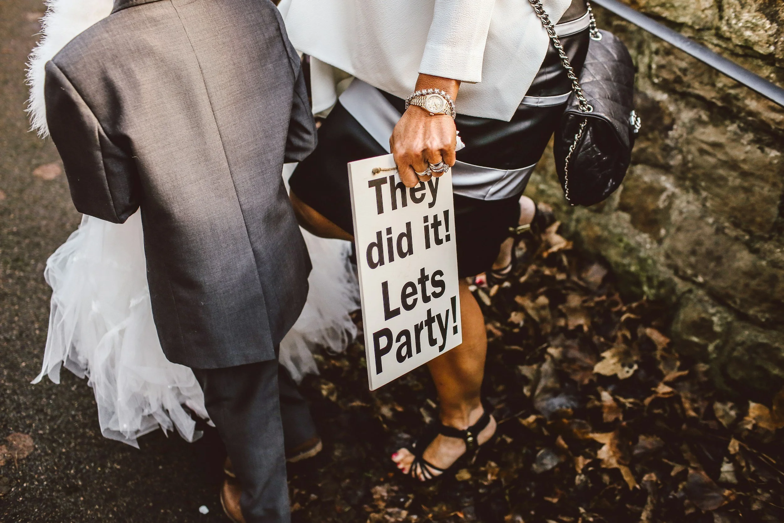 Person dressed in formal attire holding a sign that reads "They did it! Let's Party!" at an outdoor event, standing next to a person in a dress and heels on a leaf-covered ground.