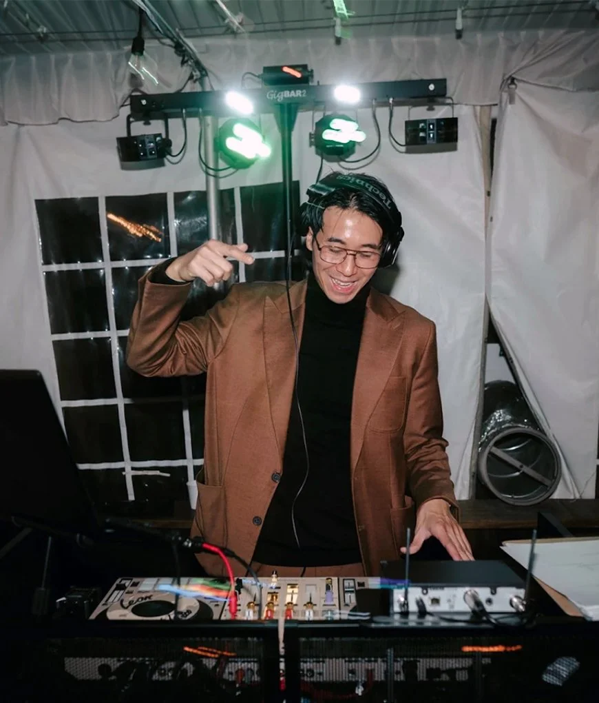 A man wearing glasses, a brown blazer, and a black shirt is DJing at a party. He is smiling, wearing headphones, and pointing to his head with one hand while operating DJ equipment. There are green lights and audio equipment behind him inside a tent 