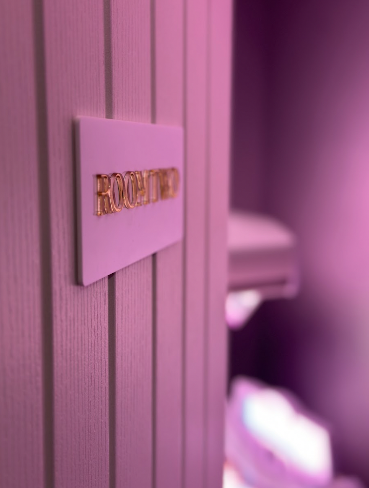 Close-up of a sign on a textured wall with the word 'Room Two' in gold letters under purple lighting.