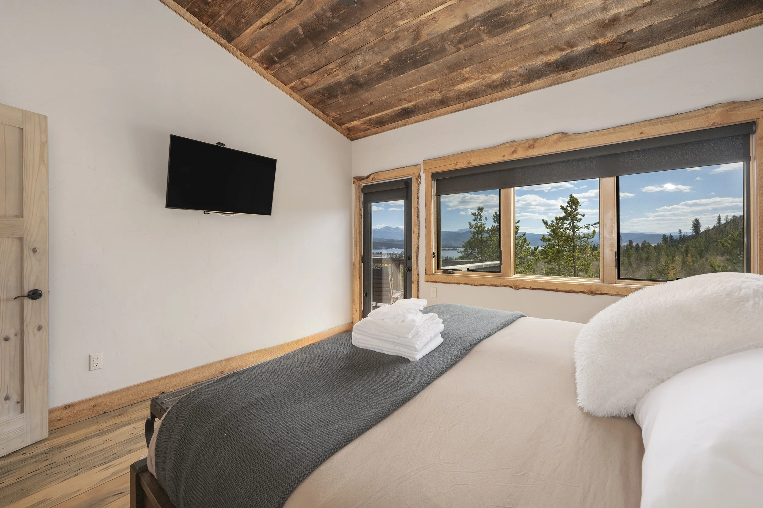 Master bedroom with a large window revealing scenic mountain and forest views, featuring an adjustable king bed with white bedding and a gray blanket, and a flat-screen TV mounted on a white wall.
