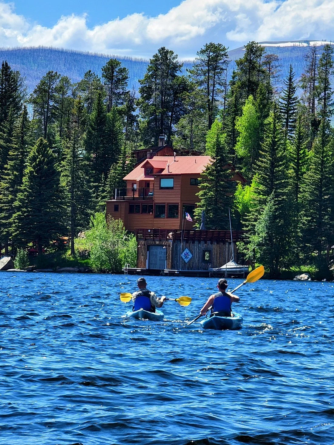Two people kayaking on a blue lake near a house surrounded by forest, with mountains in the background.