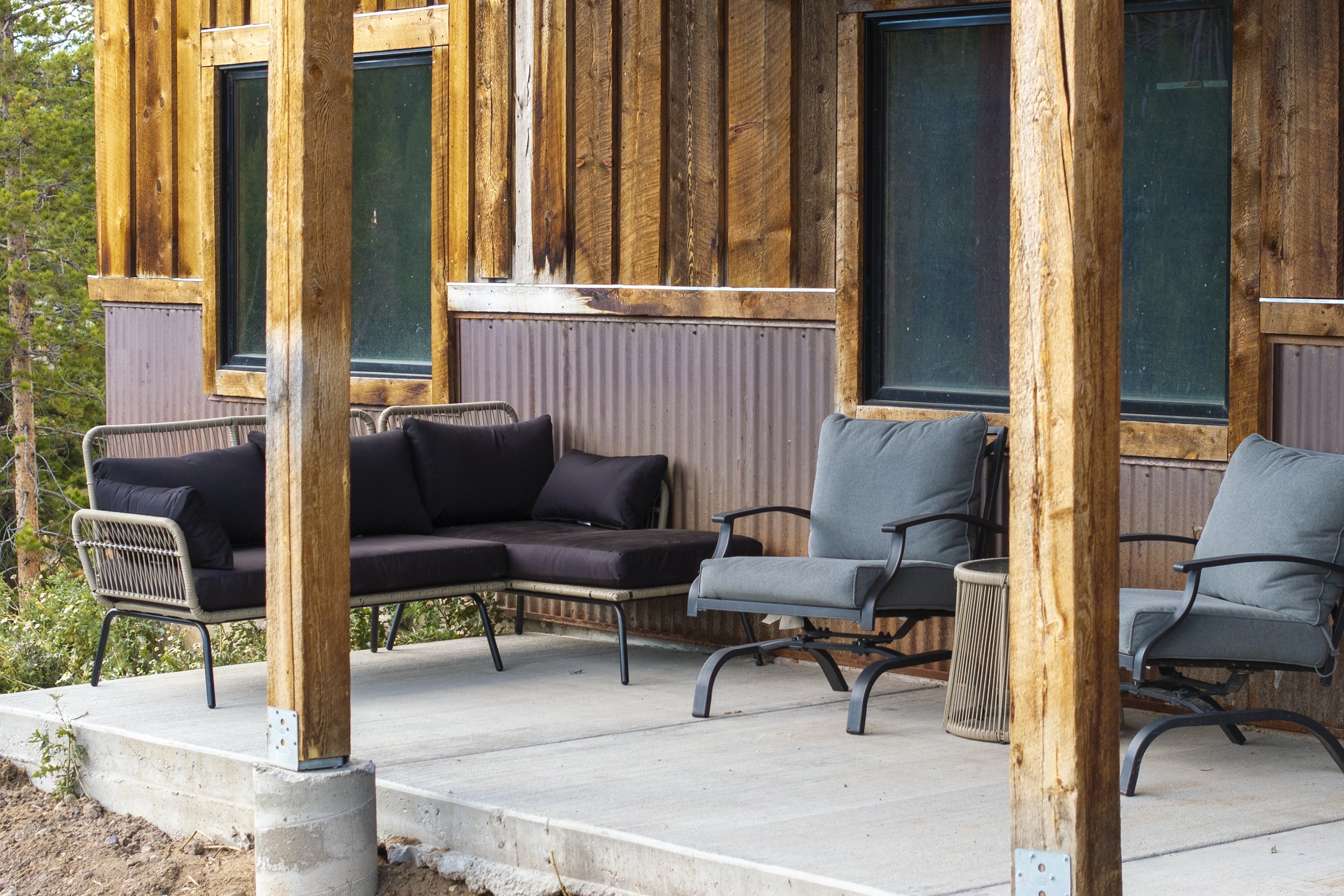 Outdoor patio with a sectional sofa and two armchairs on a concrete platform against a wooden wall with large windows, surrounded by trees.