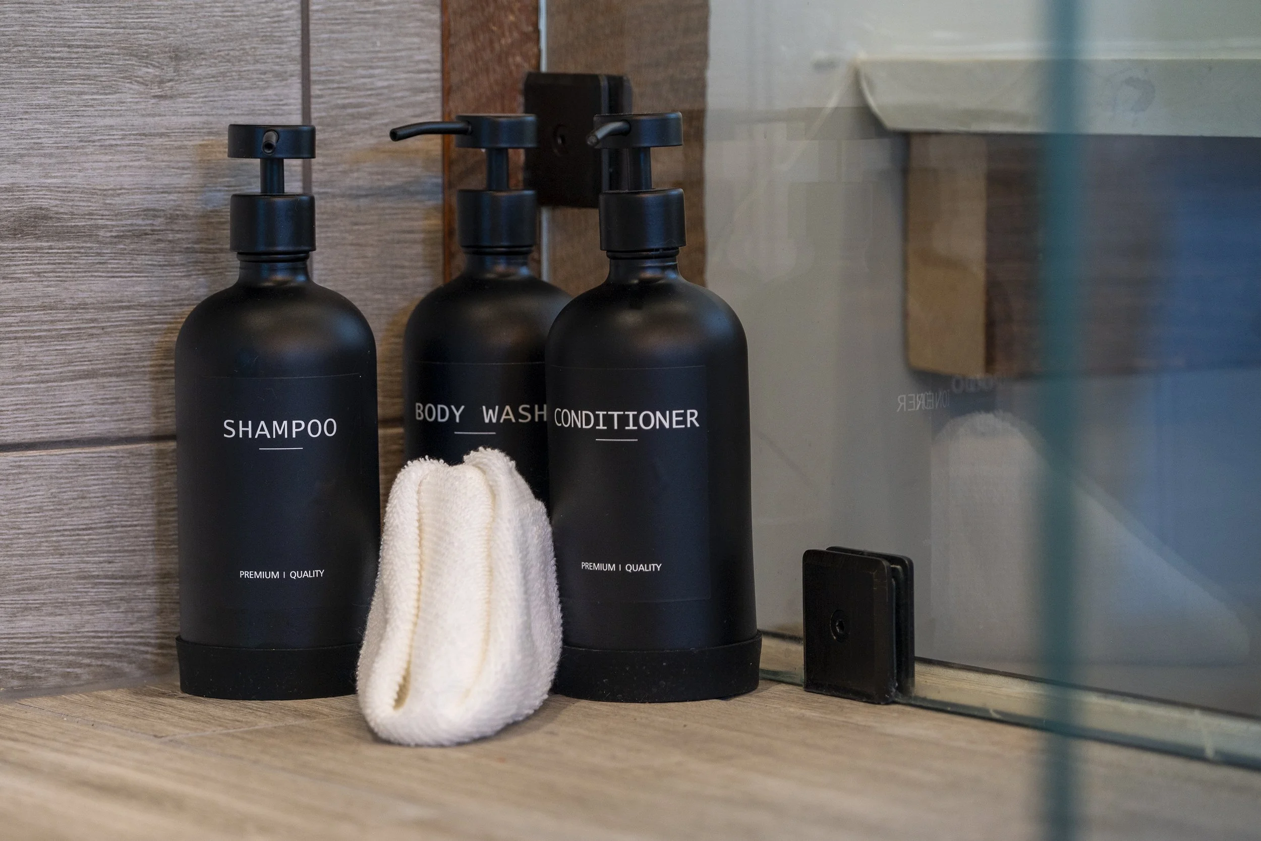 Black bottles labeled shampoo, body wash, and conditioner on a wooden bathroom shelf, with a white cloth draped over the middle bottle, next to a mirror.