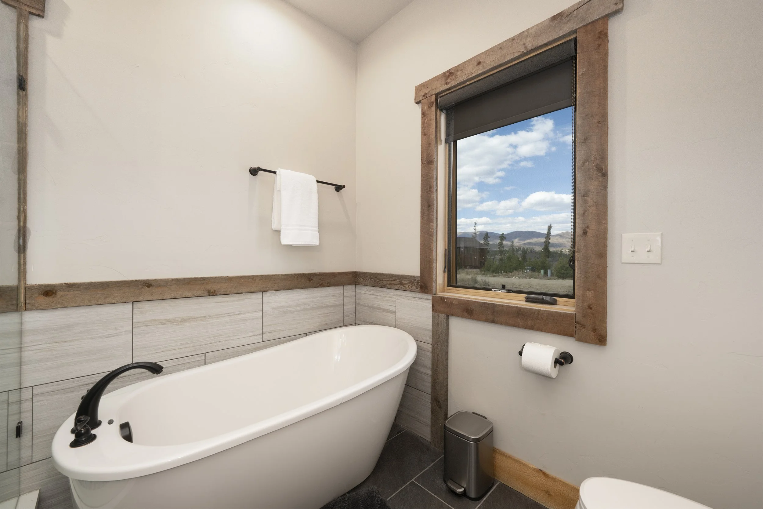 2nd master bathroom with bathtub, window showing mountain view, towel on rack, toilet paper, trash can, and rustic wooden trim.