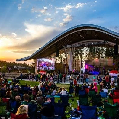 Outdoor concert at an amphitheater during sunset with a crowd seated on chairs and standing, stage with performers and colorful lighting.