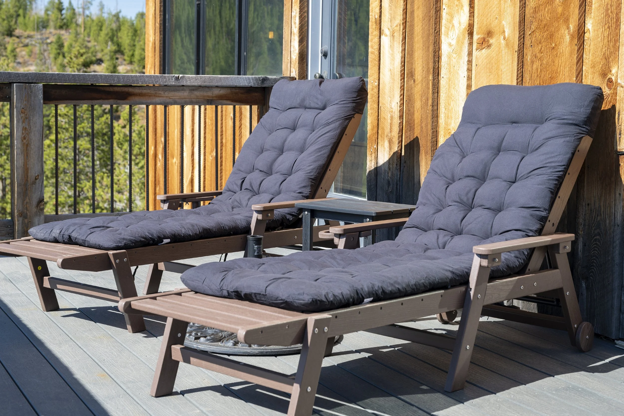 Two wooden lounge chairs with black cushions on back deck, with a view of trees and a mountain in the background.