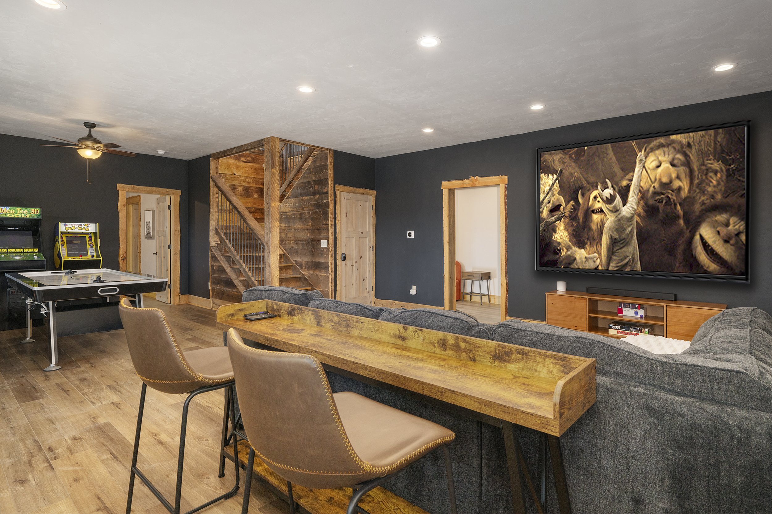 Living room with dark walls, a large porejctor screen displaying animated characters, a sectional sofa, a wooden table, arcade games, and a wooden staircase.