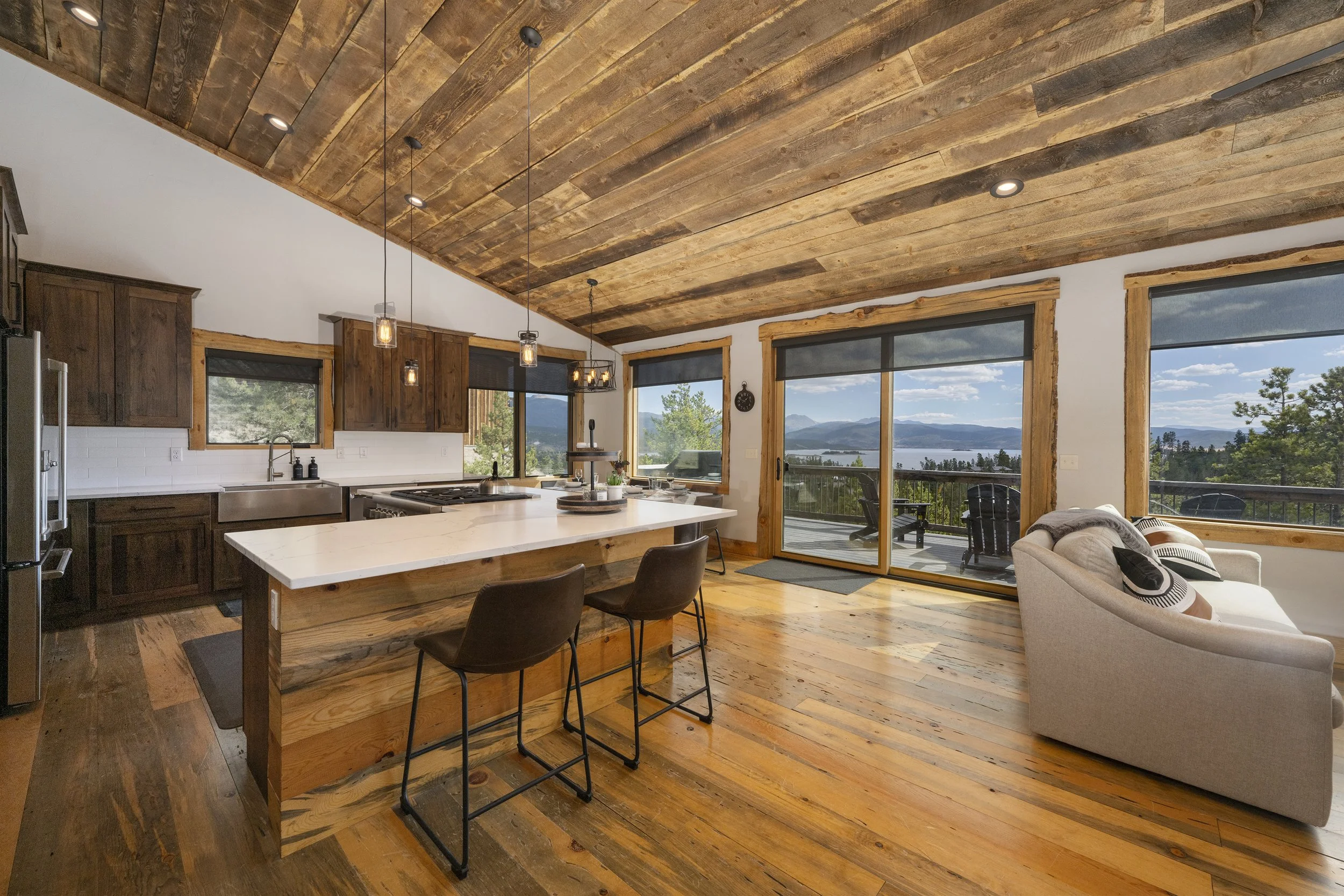 A spacious kitchen and living area with wooden floors and ceiling, featuring a kitchen island, dark wood cabinets, large windows, a sliding glass door leading to a deck with outdoor furniture, and a beige sofa with decorative pillows.
