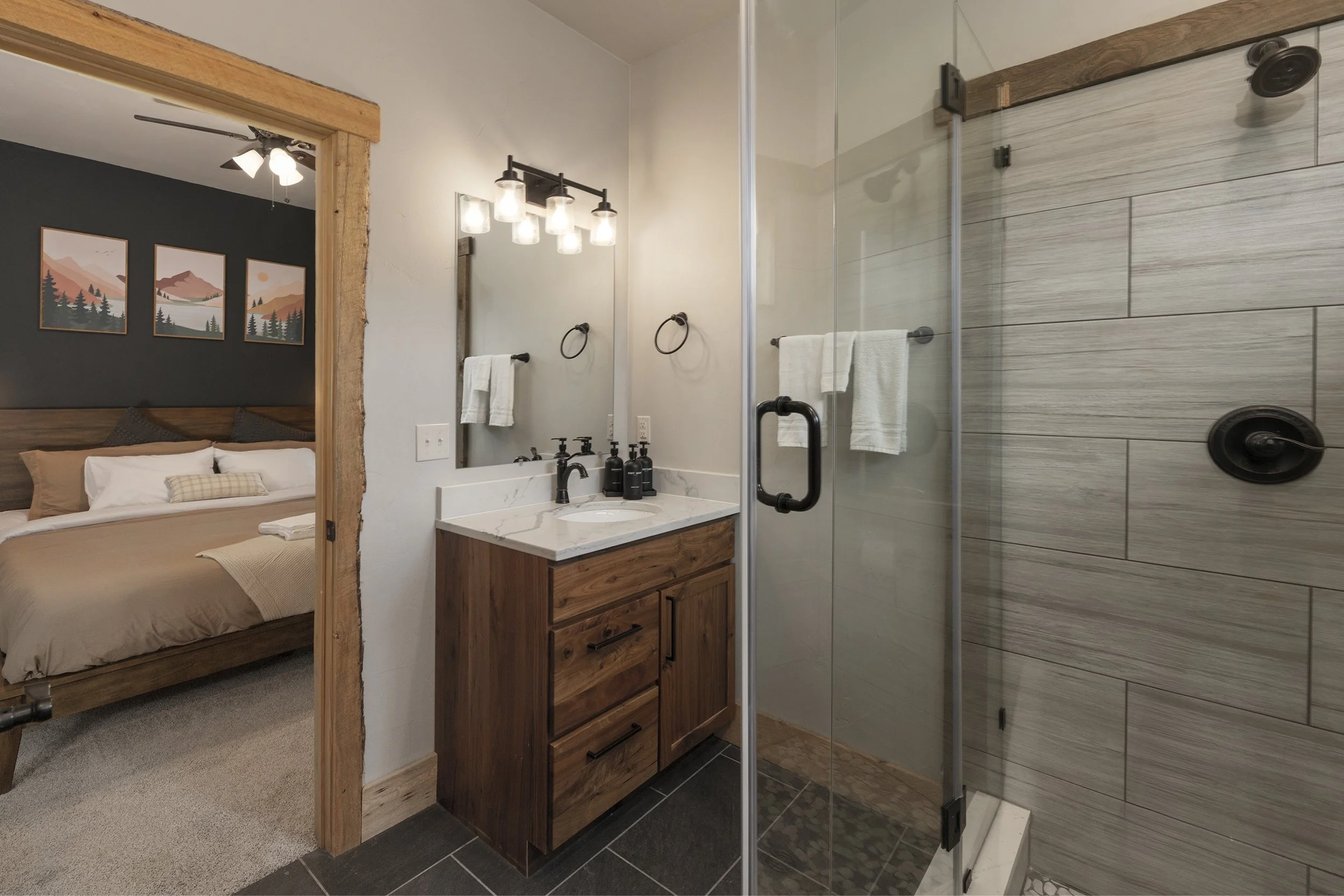 2nd master bathroom with a wooden vanity, a mirror, and a glass-enclosed shower with grey tiles. Towel bars with white towels are mounted on the walls. An adjoining bedroom with a bed, pillows, and artwork is partially visible through an open door.