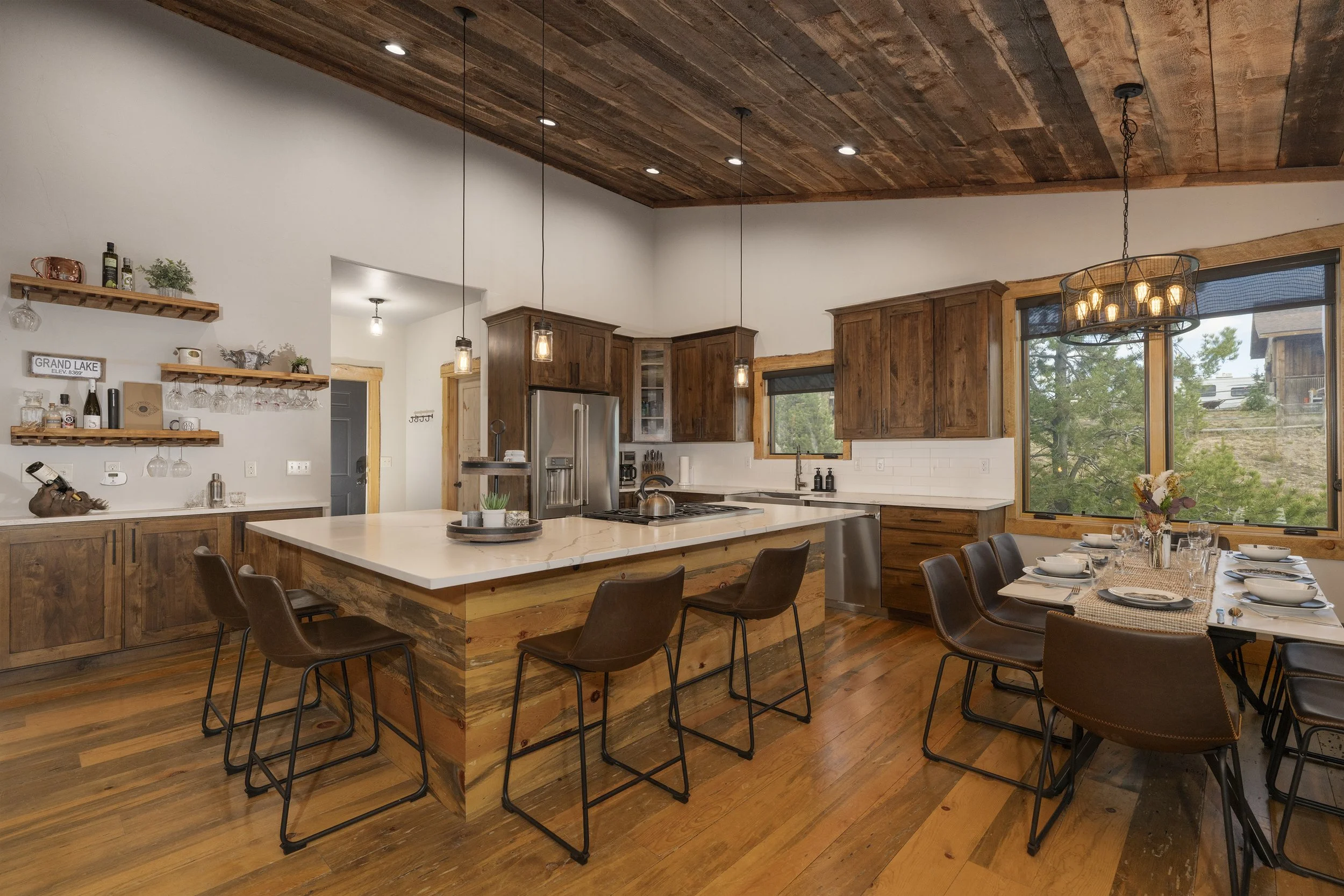 A spacious kitchen with wooden cabinets, a large island with quartz countertop, and a dining area with a table set for eight. The room features large windows with views of trees and a chandelier hanging above the dining table.