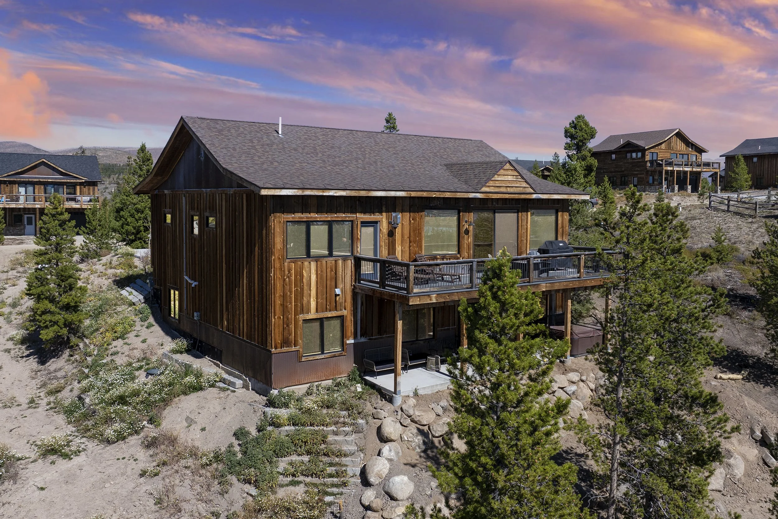 Rocky Heart Hideaway chalet exterior with deck, fire table, patio furniture, and barbecue grill, surrounded by pine trees and neighboring mountain chalets at sunset in Grand Lake, CO.