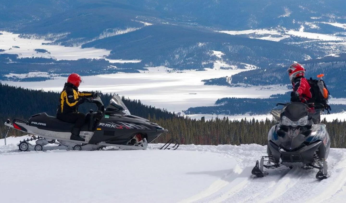Two people riding snowmobiles on a snow-covered landscape with mountains and forests in the background.