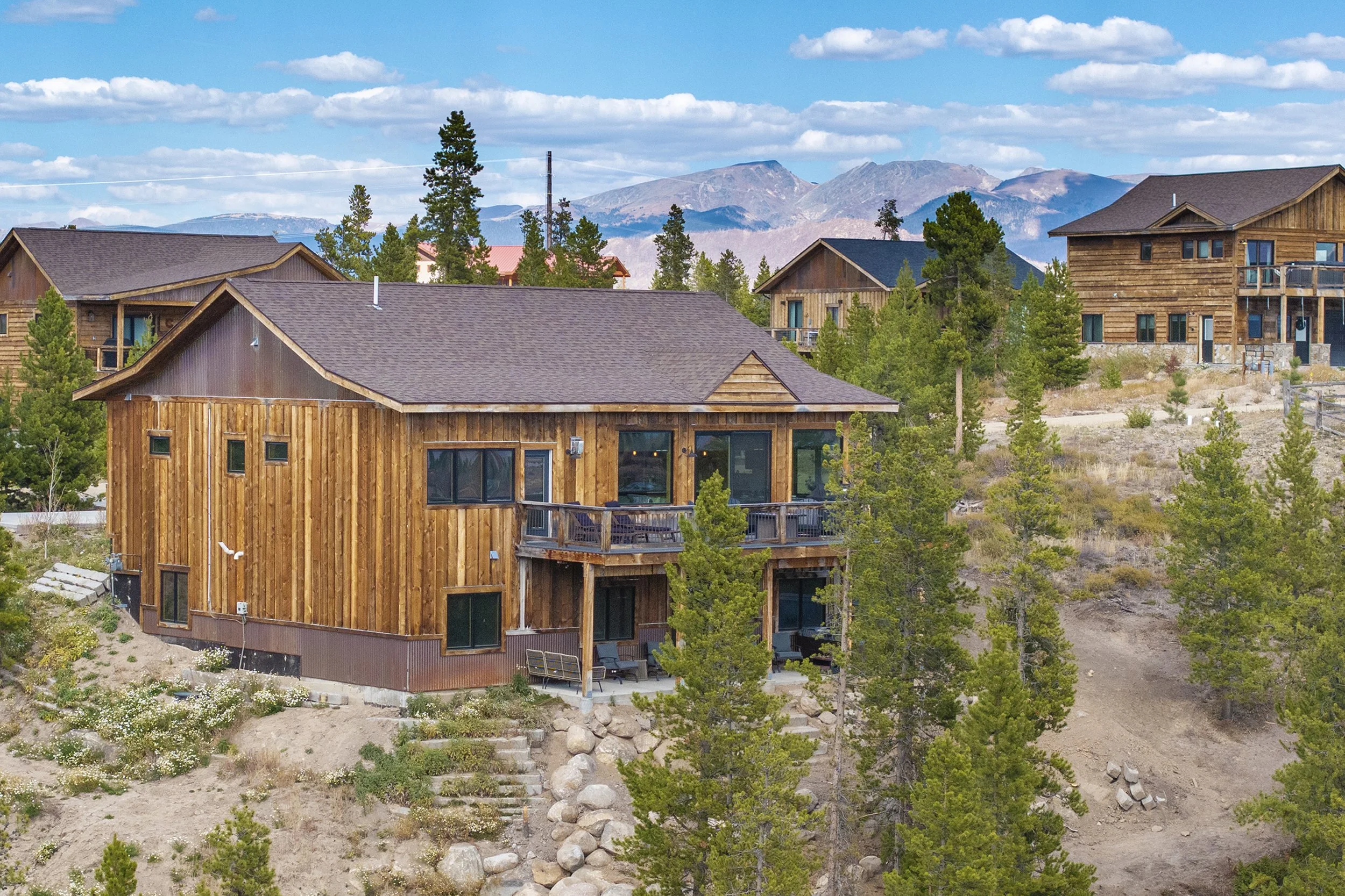 Rocky Heart Hideaway chalet on a hillside in Grand Lake, CO, with wooden exterior, surrounded by trees and mountains under a partly cloudy sky.