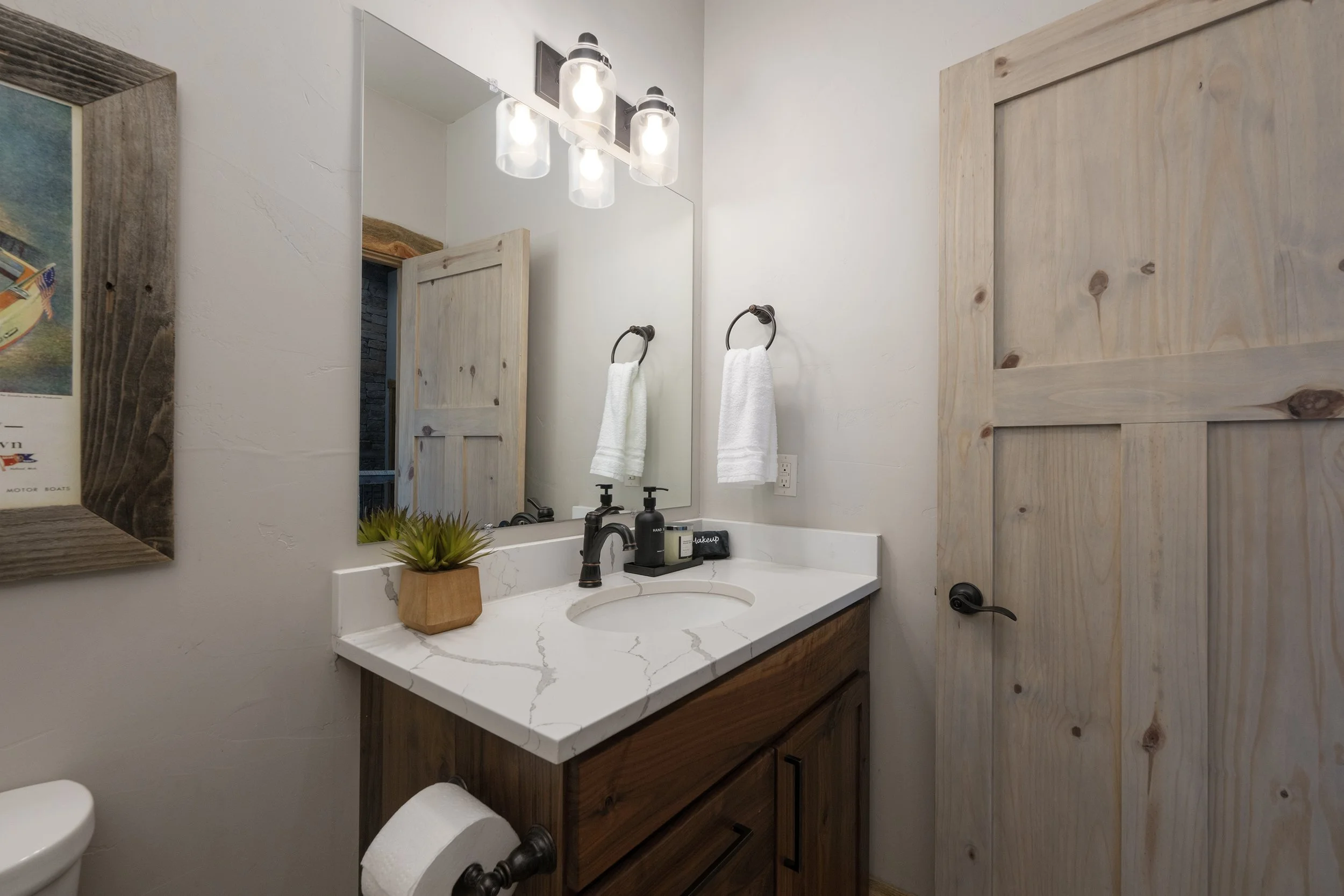 Bathroom with wooden vanity, white quartz countertop, framed mirror, black faucet, white towels, small potted plant, and wooden door.