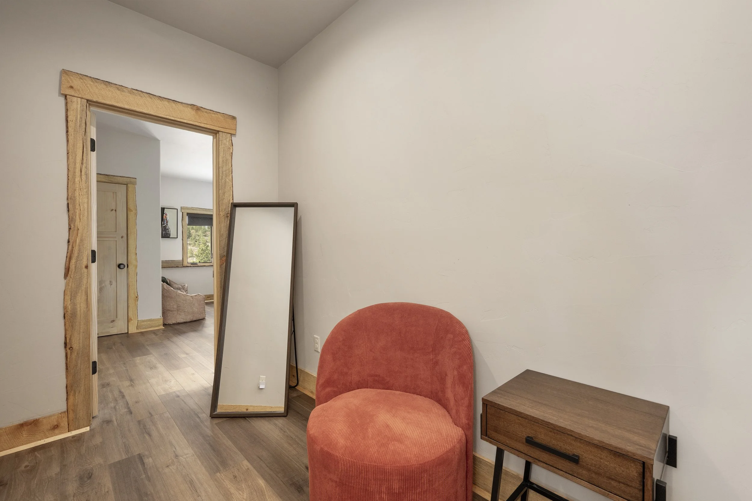 Hallway with white walls, a burnt orange upholstered chair, a wooden side table with a drawer, and a large vertical mirror leaning against the wall. There is an open doorway with wooden trim leading to the bunk room with a window and furniture.