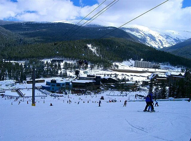 Ski resort with people skiing and snowboarding on a snow-covered slope, surrounded by trees and mountains with ski lifts overhead.