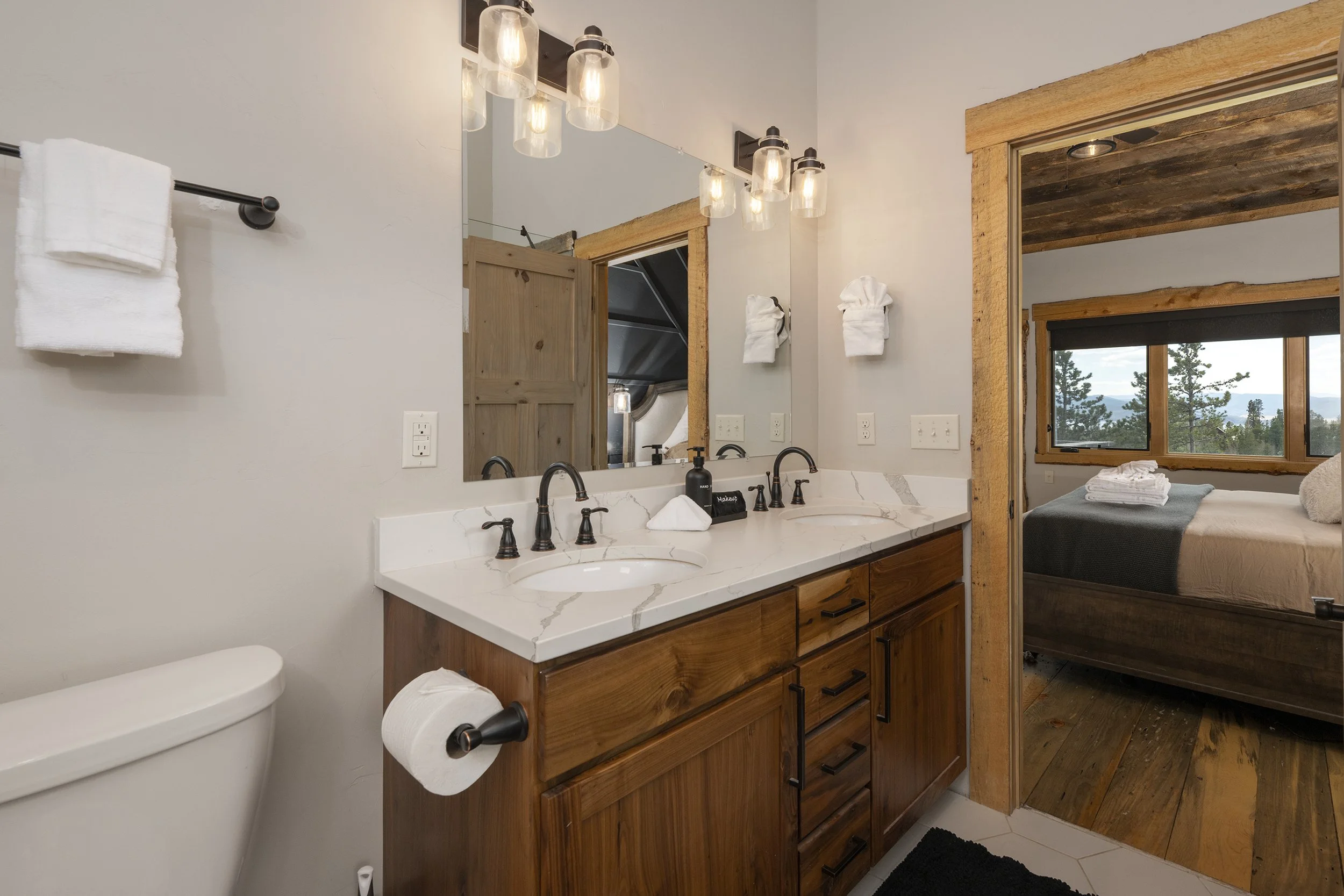 Master bathroom with double vanity sink, large mirror, stained wood cabinetry, wall-mounted towel rack with towel, toilet, and view of bedroom with bed and window showing trees and mountains outside.