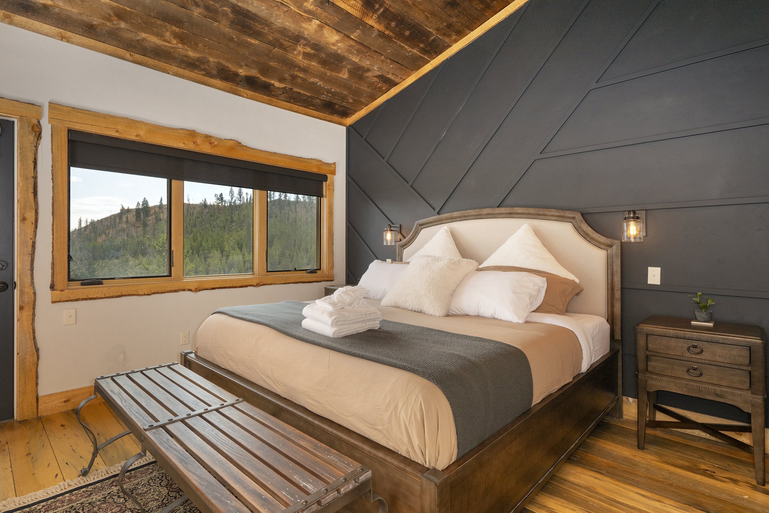 Master bedroom with a large adjustable king bed, wooden bench at the foot of the bed, and a 2 nightstands. The room has wooden floors, a window with a mountain view, and a dark paneled accent wall behind the bed.