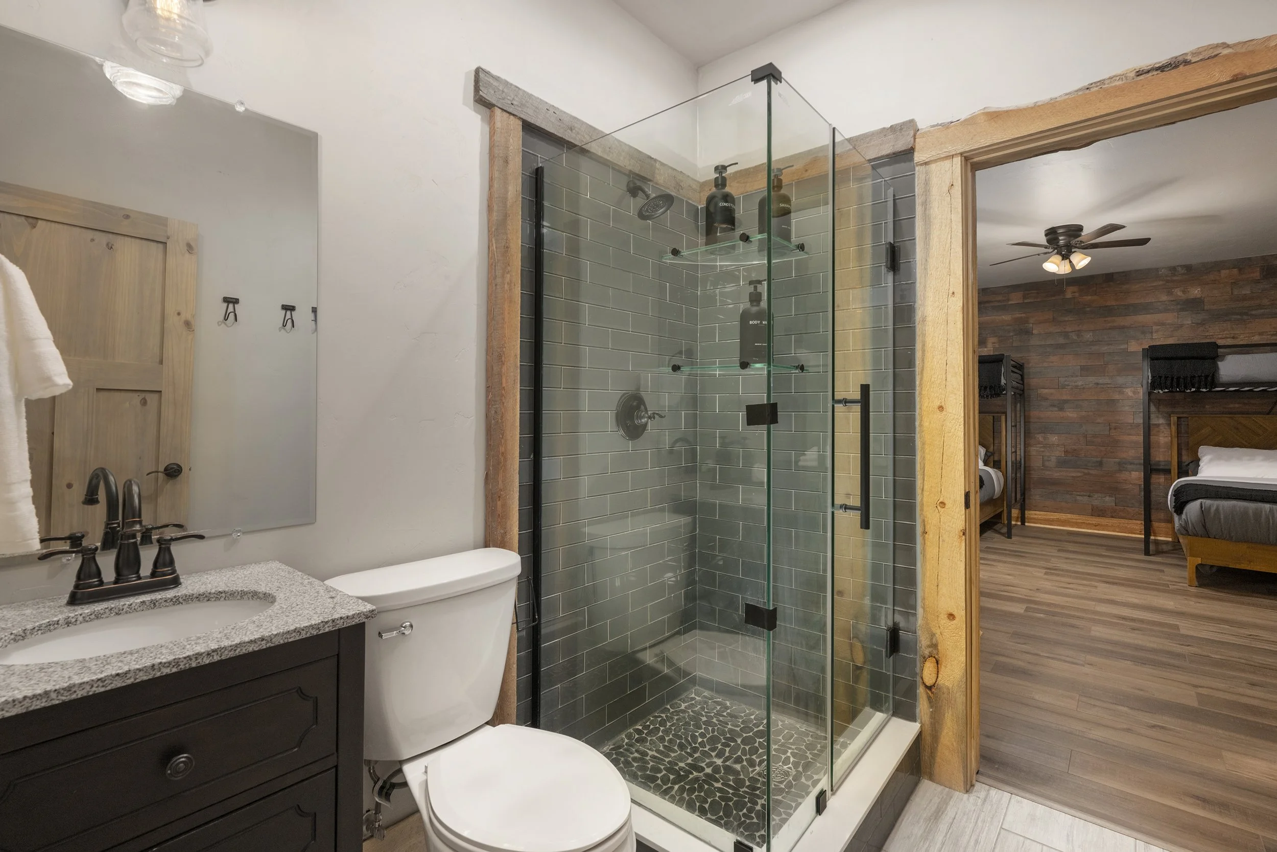 Basement bathroom with a glass shower, a toilet, a dark wooden vanity with a quartz countertop, and a mirror, adjacent to the bunk bedroom with wood-paneled walls and floors, ceiling fan, and beds.