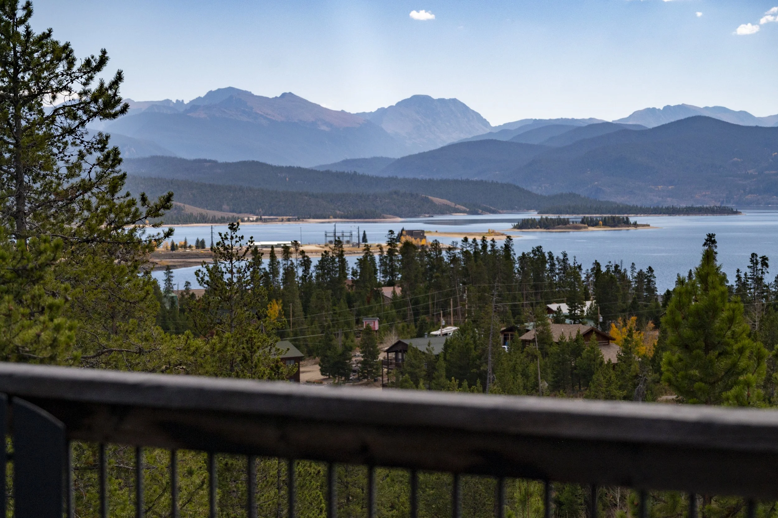 Scenic view from Rocky Heart Hideaway chalet in Grand Lake, CO, overlooking the lake, forested hills, and mountains, with a wooden railing in the foreground.