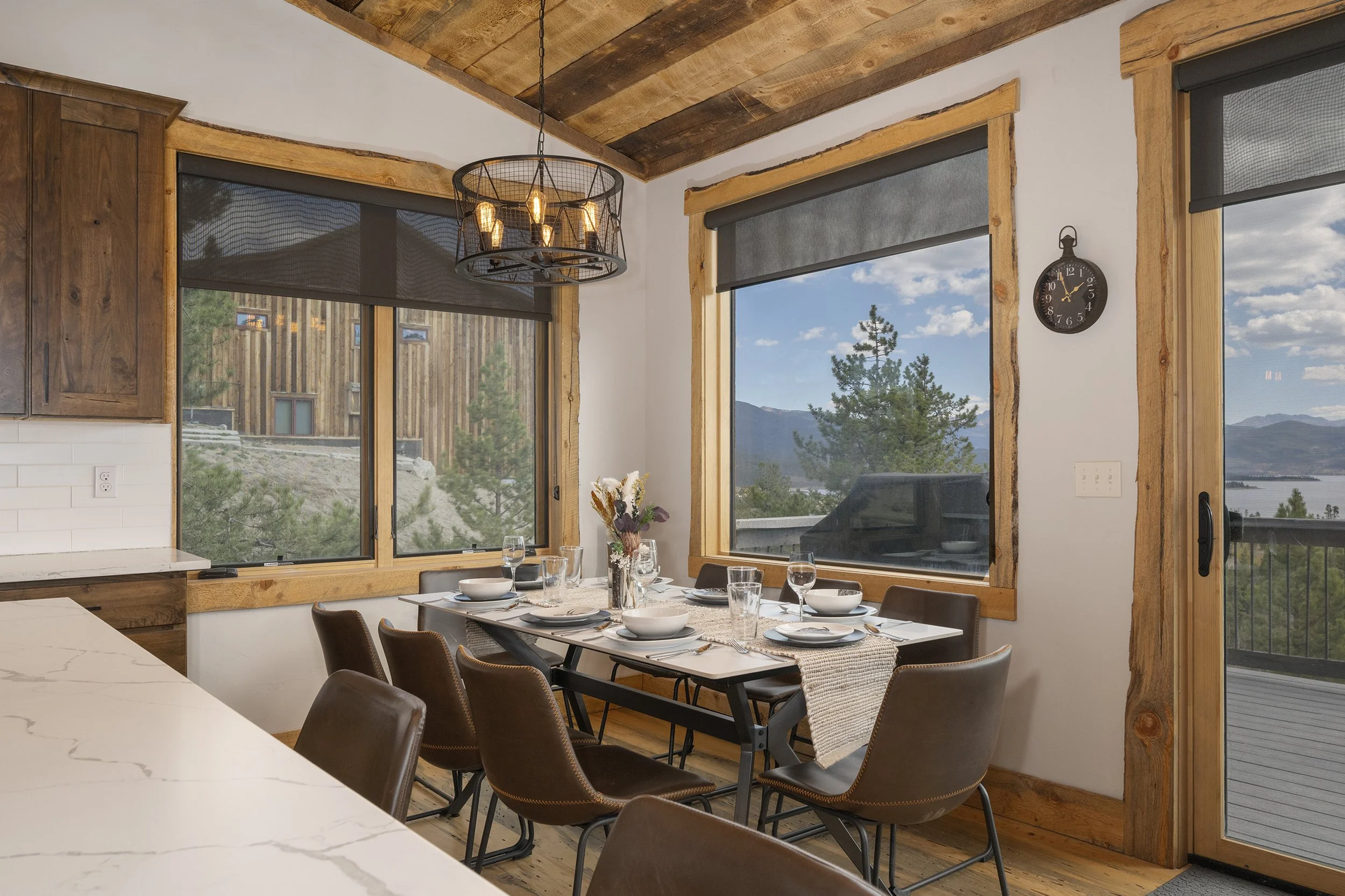 Dining area with a table set with white plates, glasses, and a vase of dried flowers, surrounded by brown chairs, large windows showing mountain and lake views, wooden ceiling and trim, and a rustic clock on the wall.