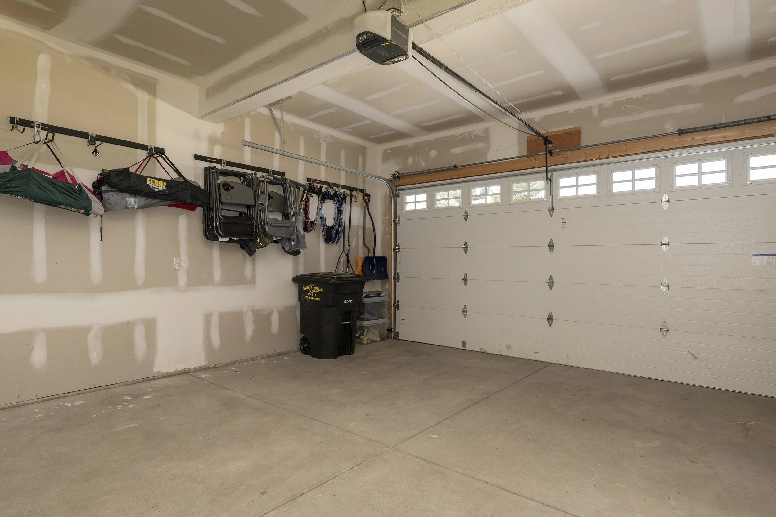 2 car garage with a closed white garage door, storage hooks with lawn chairs, snowshoes, and and garabge can.