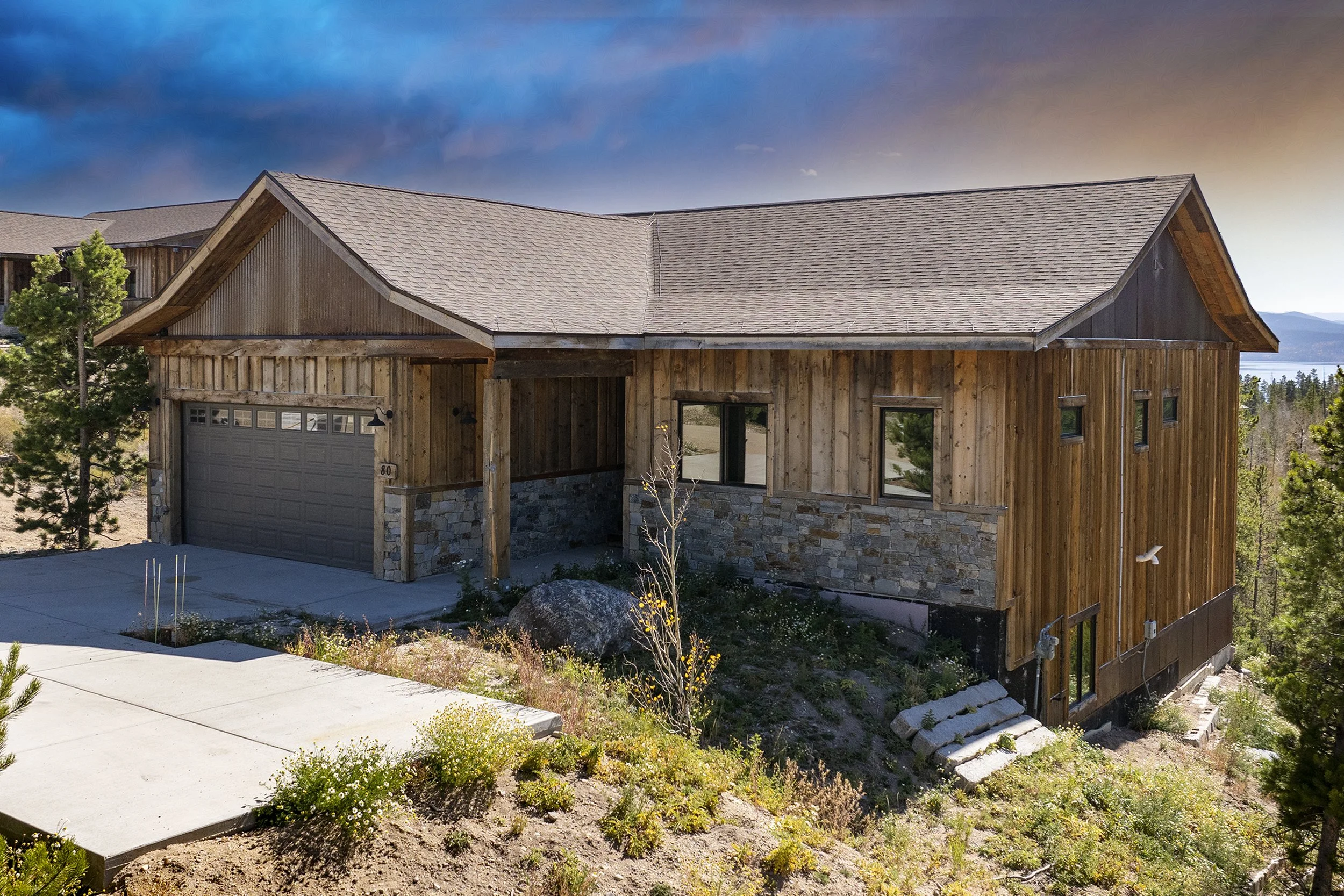 Exterior of Rocky Heart Hideaway chalet in Grand Lake, CO, featuring rustic wooden and stone design, pitched roof, attached garage, sloped terrain with plants and rocks, surrounded by trees and mountains under a partly cloudy sky.