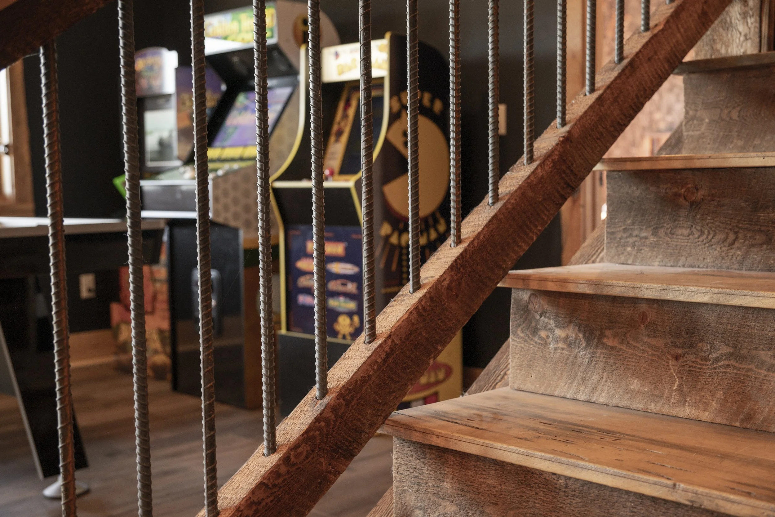 Close-up view of a wooden staircase with metal railing spindles leading to a basement arcade area with arcade machines.
