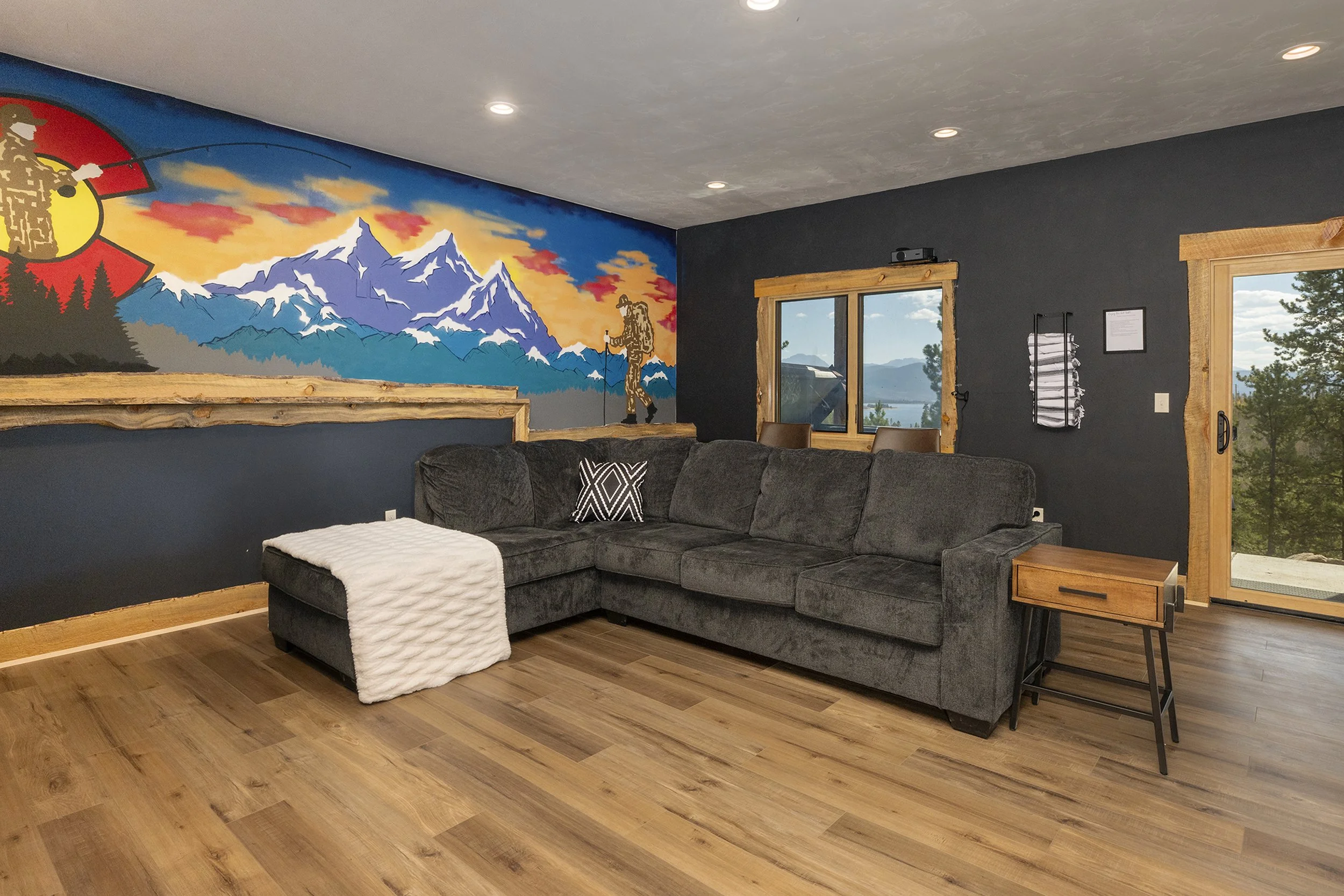Living room with dark gray sectional sofa, wooden framed windows, mountain mural, and scenic view of lake and trees outside.