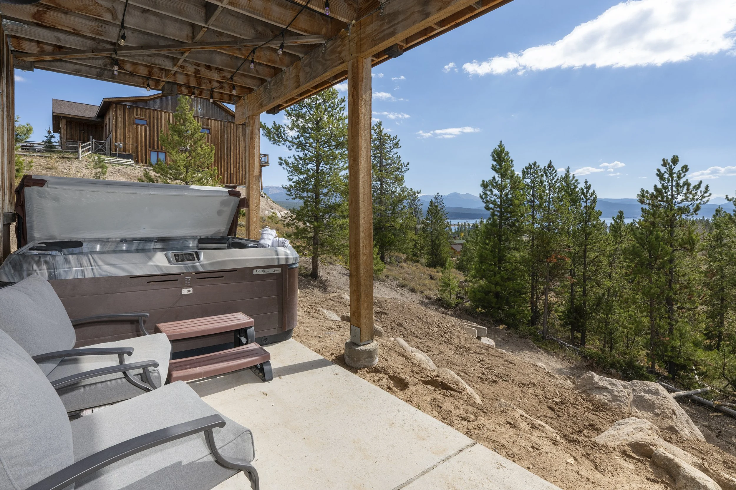 Outdoor patio with hot tub, cushioned chairs, and a small bench, overlooking a wooded landscape with trees, mountains, and a lake in the distance under a blue sky.