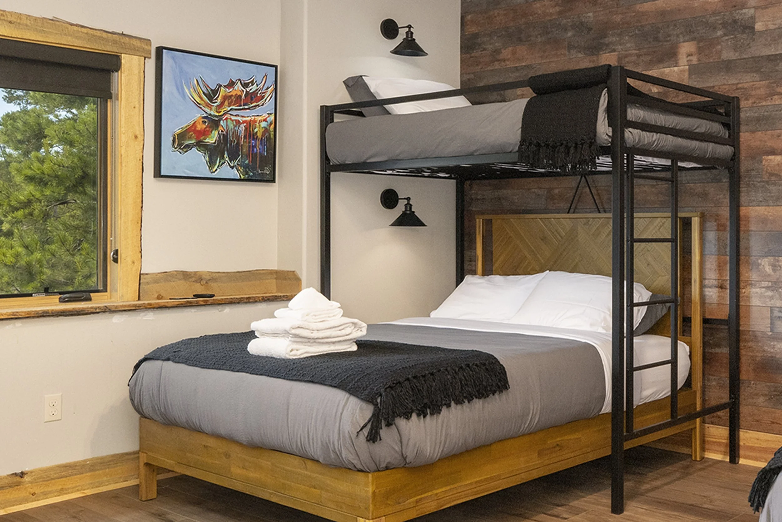 Basement bunk bed room. Bottom is a queen size bed and the top is a twin size bed, featuring wood and black metal frames, a wooden headboard, a window with wooden trim, a colorful moose artwork, and a black wall-mounted lamps.