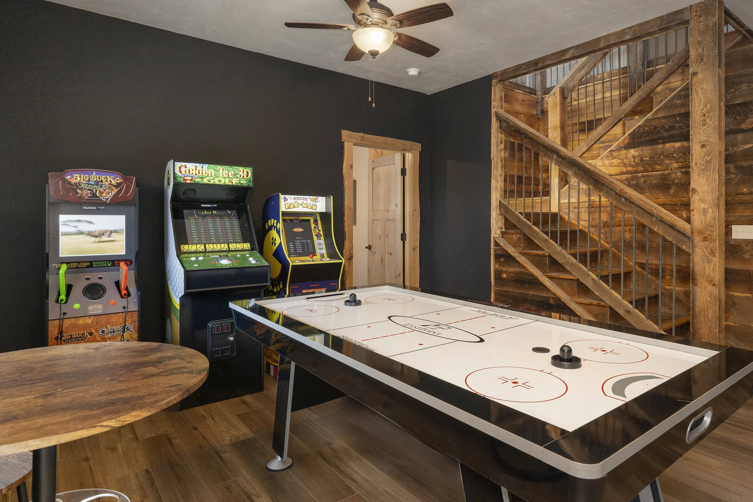 Indoor recreational room, wooden staircase, and an air hockey table in the center. There are three vintage arcade games against the wall on the left side.