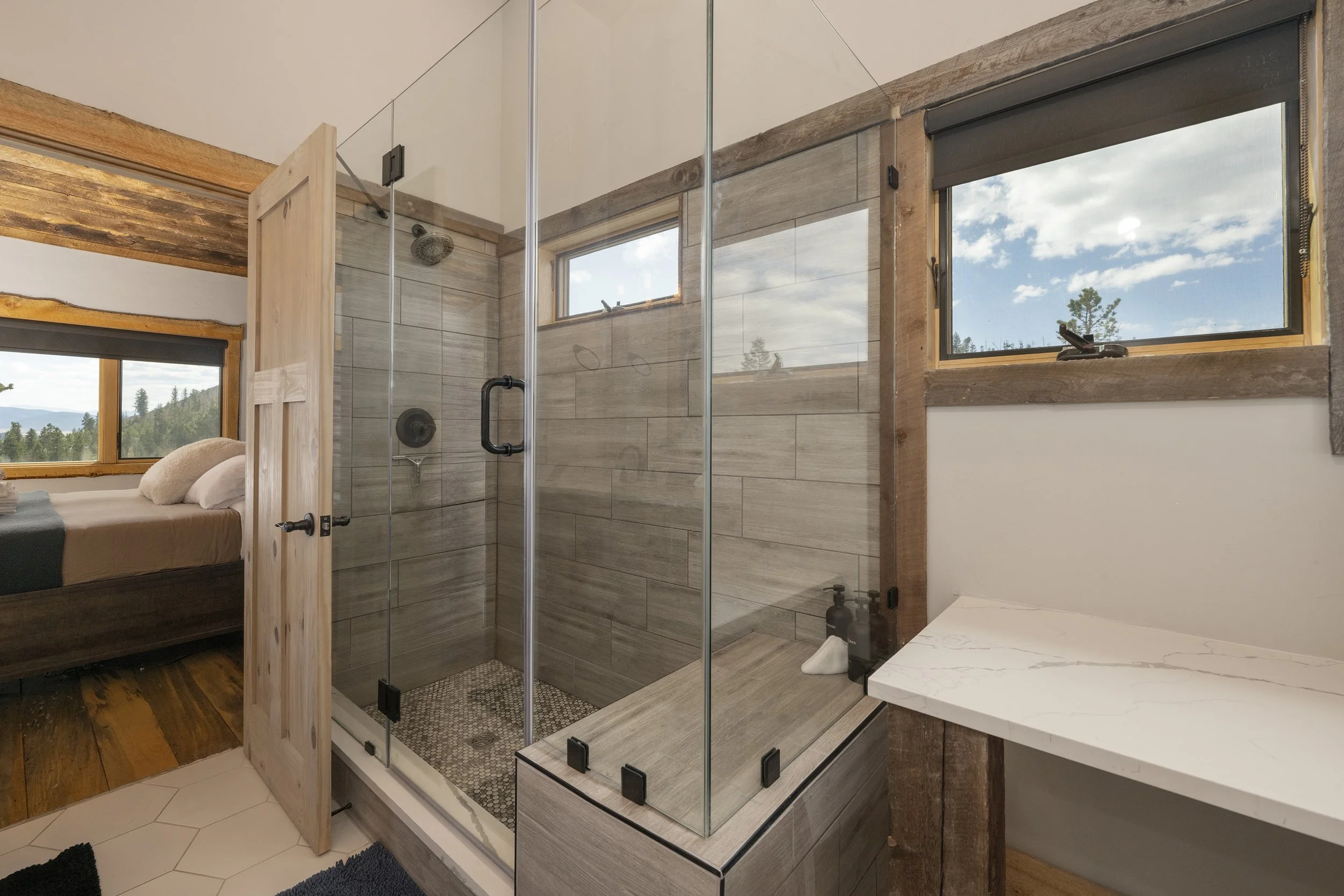 Master bathroom with a glass shower enclosure, wood accents, and two horizontal windows showing a scenic outdoor view.