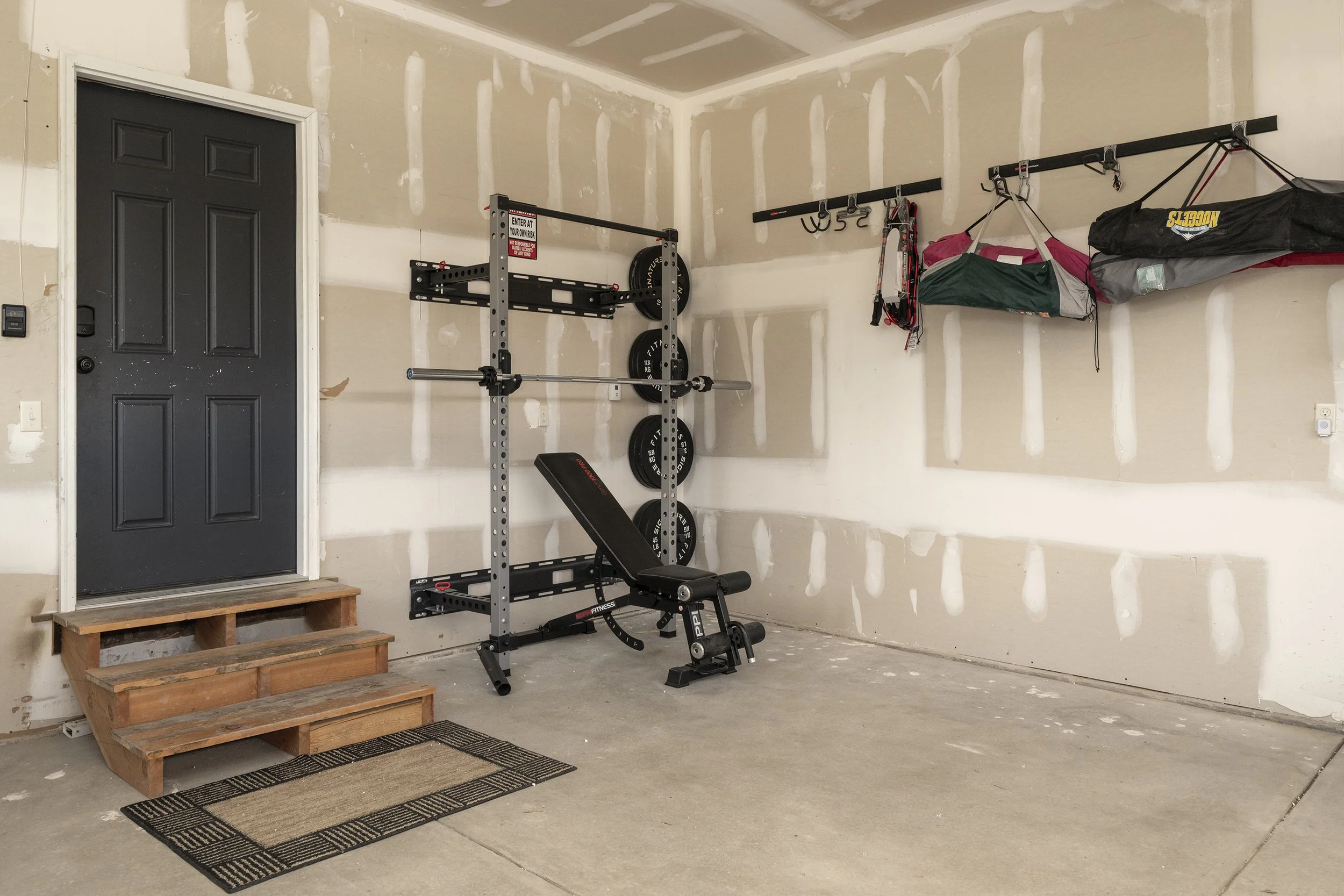 A garage with a black door, a small wooden staircase, and a workout station including a bench, weights, and a pull-up bar, along with storage hooks on the wall holding lawn chairs.