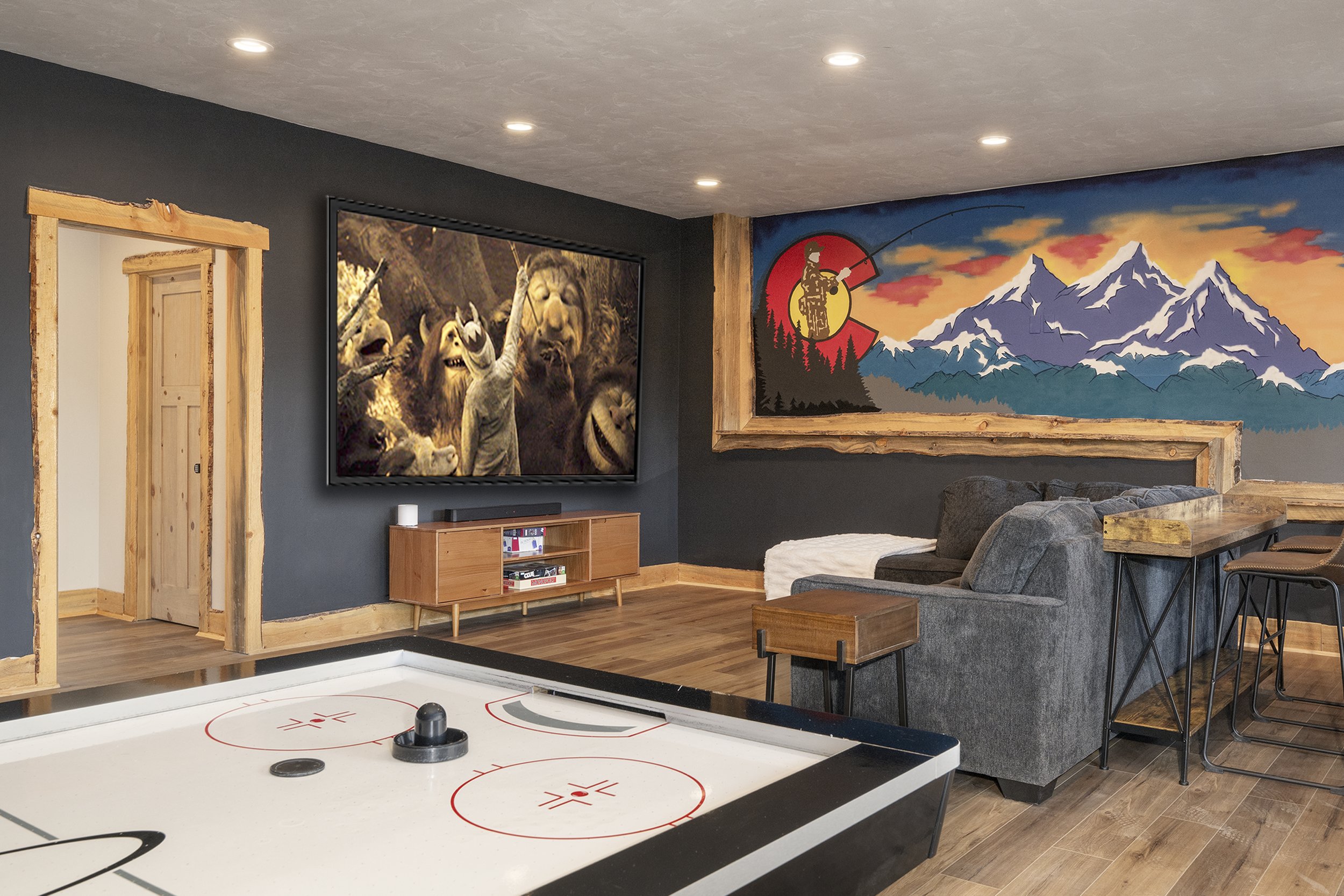 A recreation room with a air hockey table, a large wall art mural of mountains, a projection screen displaying animated characters, a wooden door frame, and a gray couch.