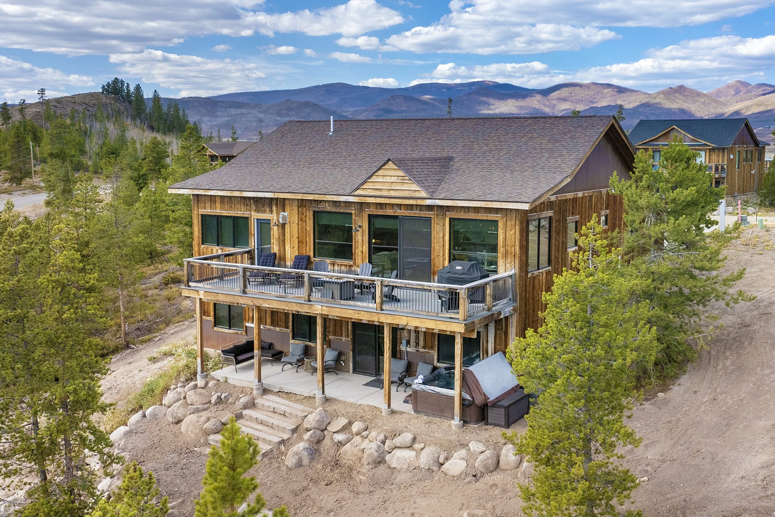 Two-story Rocky Heart Hideaway chalet in Grand Lake, CO, featuring wooden exterior, deck, large windows, and surrounded by trees with mountains in the background.