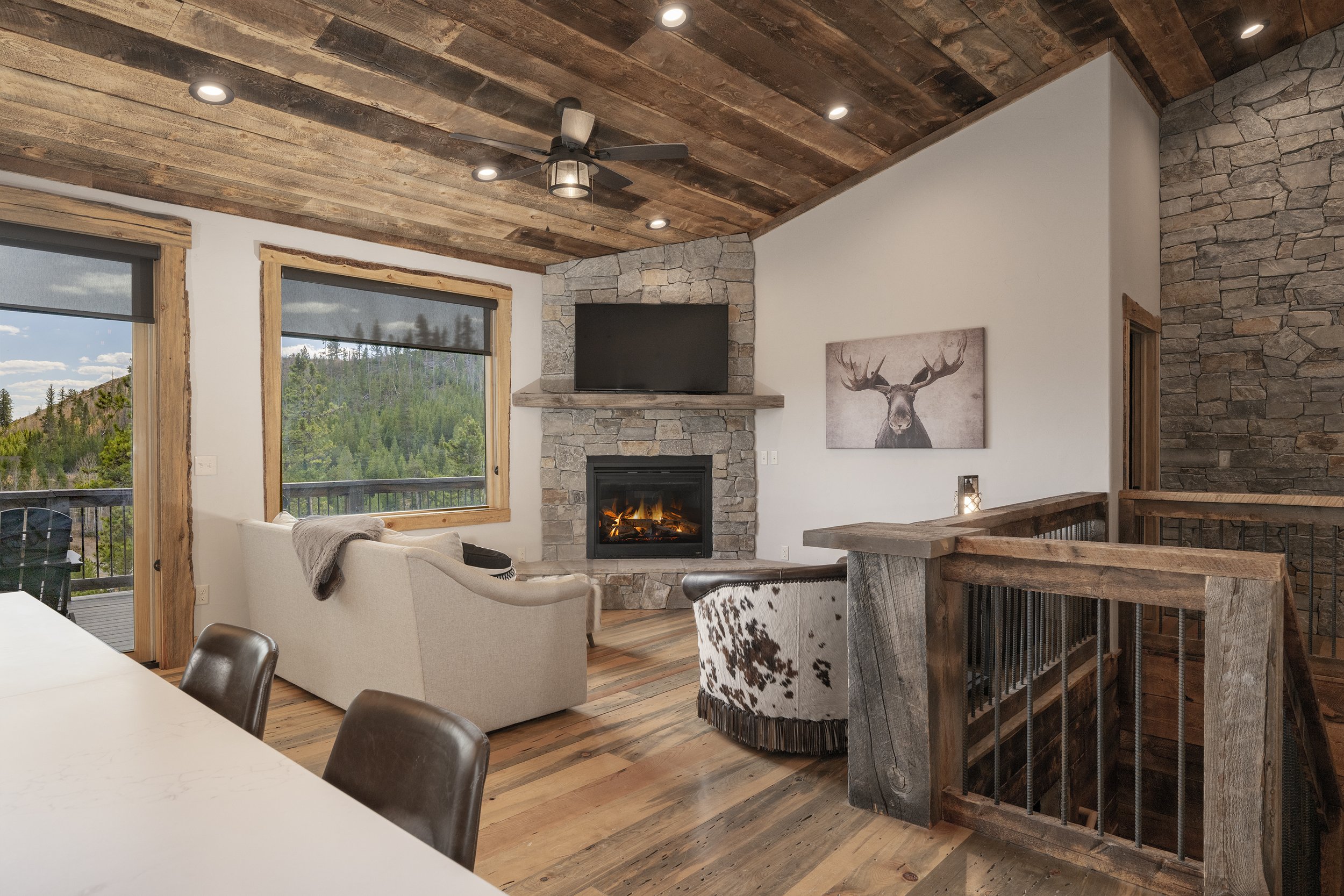 Living room with wooden ceiling, stone fireplace with TV mounted above, large windows showing a mountain view, a white sofa, a cowhide chair, and wood railings.