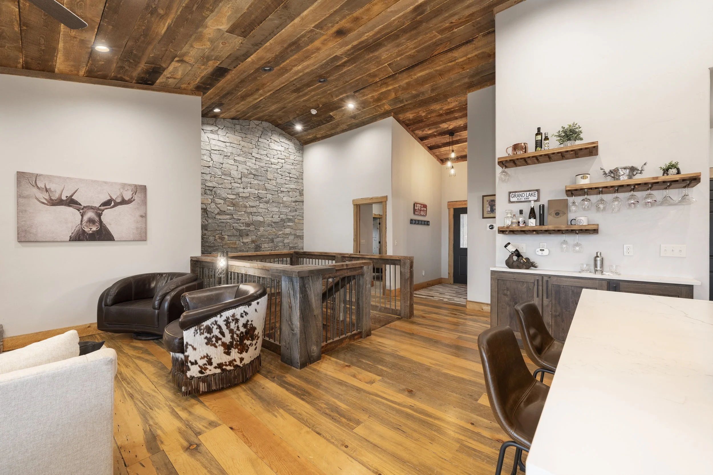 Modern living space with wood-paneled ceiling, stone wall, wooden flooring, and furniture including a leather armchair, cowhide chair, and a white couch. Decor includes artwork of a moose, wall-mounted shelves with bottles and glassware, and a black front door.