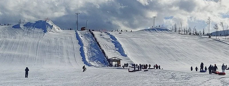 People at a snowy ski jumping hill with a steep ramp leading to a platform, surrounded by snow and cloudy skies.