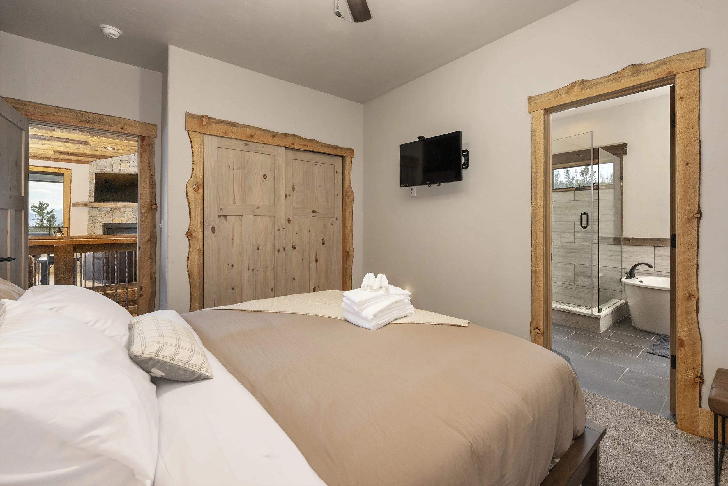 2nd master bedroom with a king bed, folded towels, and a bathroom visible through a door. Wooden accents around the doorway, small wall-mounted TV, and a view of a living room with a fireplace and window in the background.