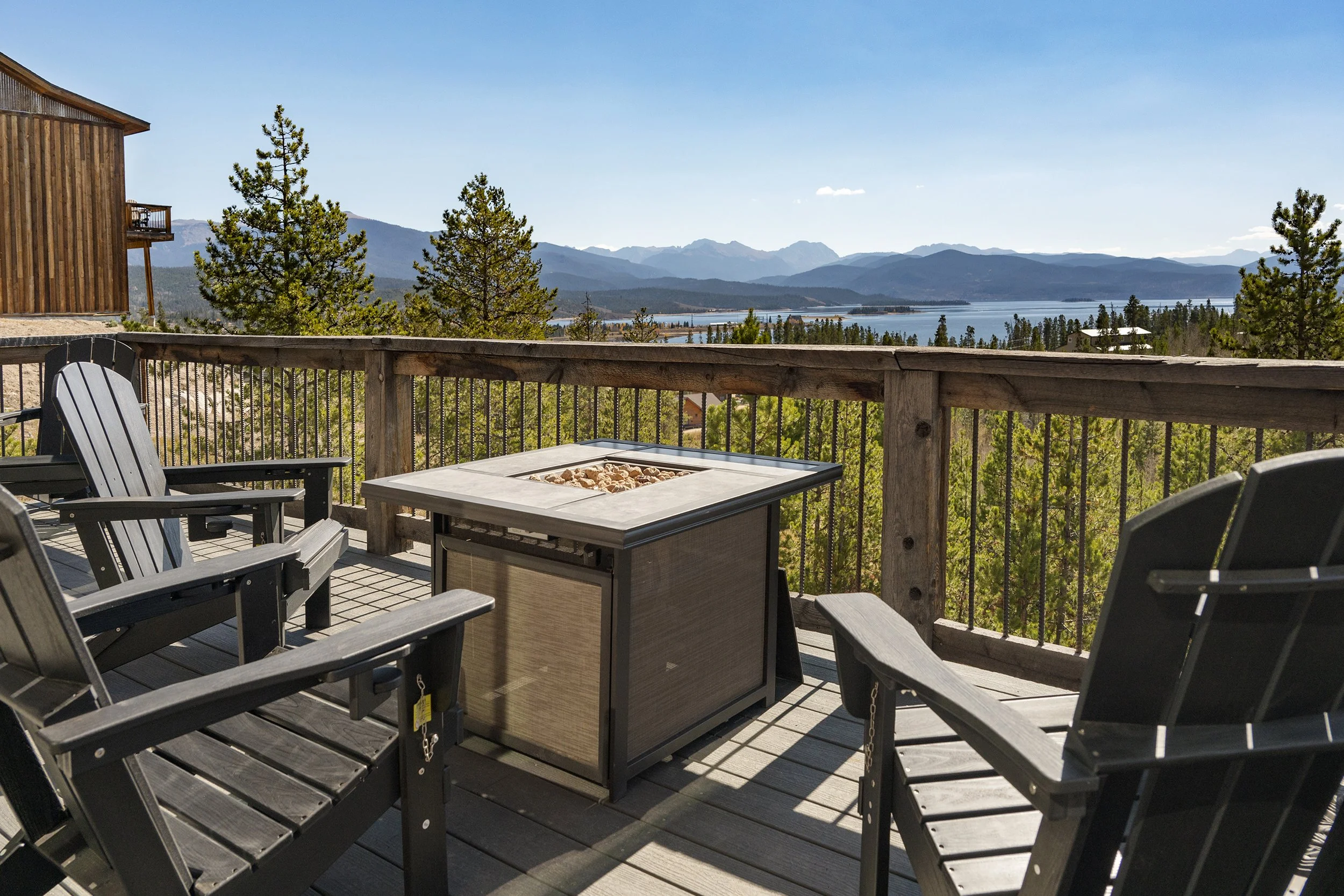 Back deck at Rocky Heart Hideaway chalet in Grand Lake, CO, with black chairs and fire pit table overlooking the lake, pine trees, and mountains under a clear blue sky.