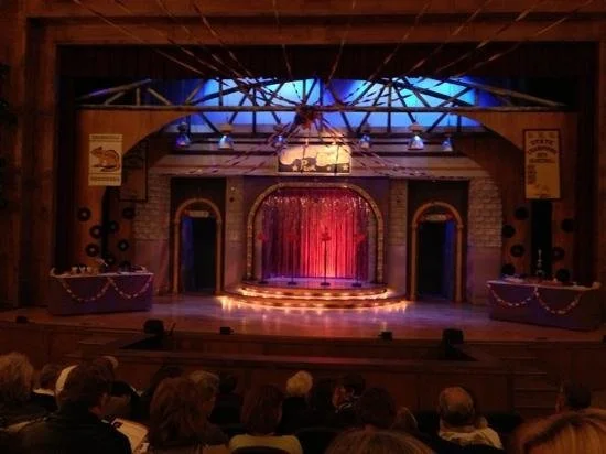 Stage with red curtains, decorated with string lights, and two side tables, in a theater with audience watching.