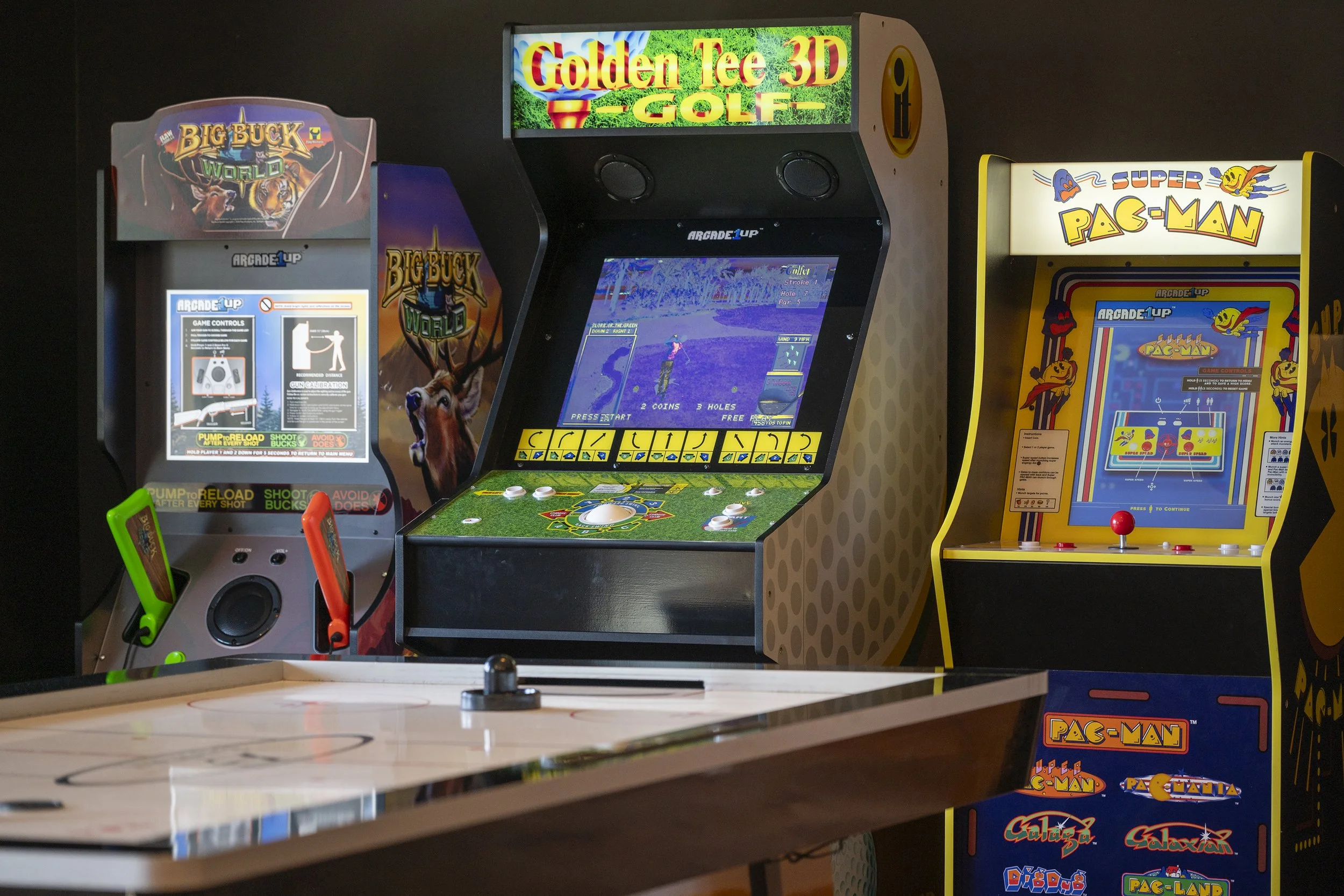 Three arcade game machines in a row, featuring Big Buck World, Golden Tee 3D Golf, and Super Pac-Man, with a portion of an air hockey table in the foreground.